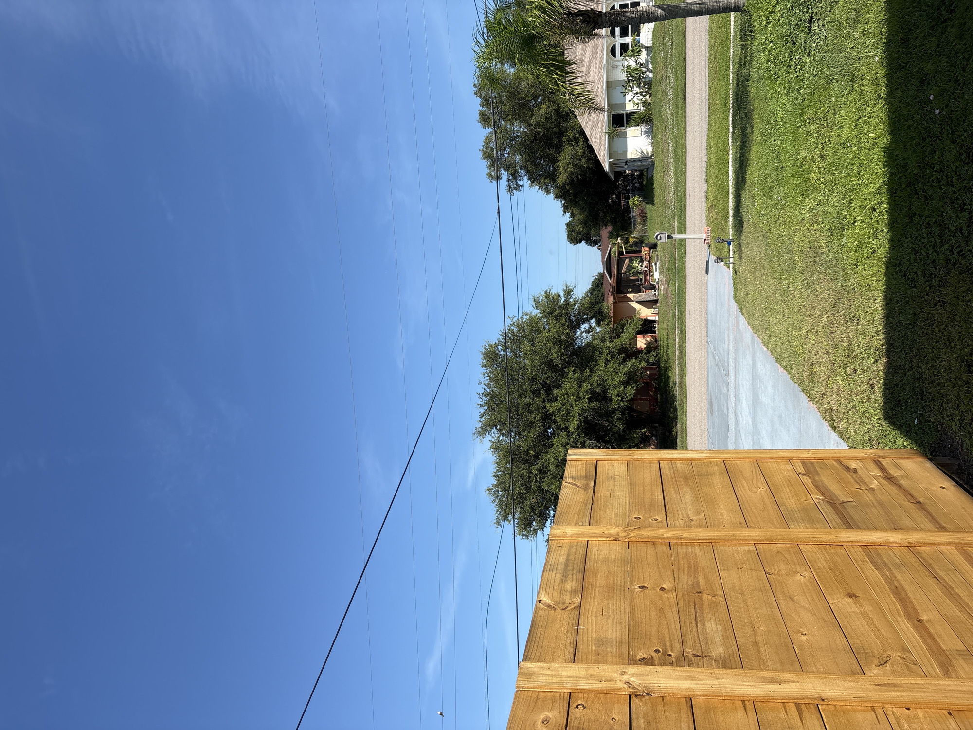 This image depicts a residential street scene on a clear, sunny day in North Port, USA. The perspective is from behind a newly constructed wooden fence, looking upwards and to the side. The bright blue sky dominates the upper left portion of the frame, with a few wispy clouds scattered across it. Several parallel power lines stretch across the sky, casting faint shadows.
To the right, the scene transitions to a well-maintained residential area. A paved driveway runs alongside a lush green lawn. In the background, houses with typical suburban architecture are visible, some with palm trees and other landscaping. One house in the mid-ground features a prominent A-frame roof and white siding, with arched windows. Another house, further back and to the left, has a more muted color scheme with visible foliage around it.
The most prominent object in the foreground is the wooden fence, constructed of vertical planks with horizontal support beams. Its warm, natural wood tone suggests it is recently built or stained. The overall atmosphere is peaceful and idyllic, characteristic of a suburban environment on a pleasant day. The time of day appears to be midday, given the bright sunlight and the quality of the shadows cast. No people or animals are visible, and there is no visible text in the image.