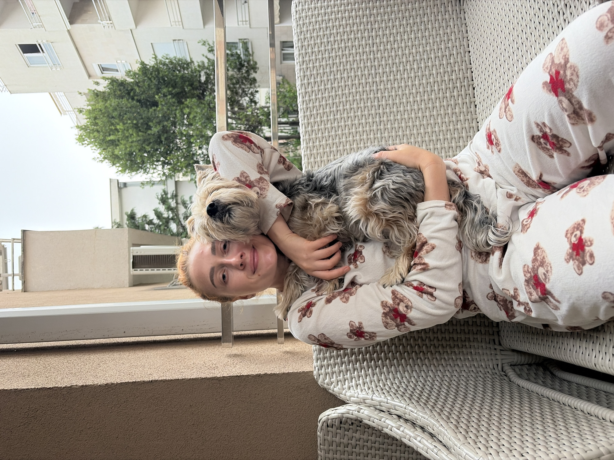A Caucasian woman in teddy-bear-printed pajamas is reclined in a white wicker chair on a balcony, cradling a small, shaggy dog with gray and tan fur. The balcony has a light brown concrete wall, and a metal railing is visible in the background. The scene overlooks a building with white balconies and a green tree. Based on the architectural style and vegetation, the location is likely in the city of Budva, Montenegro.