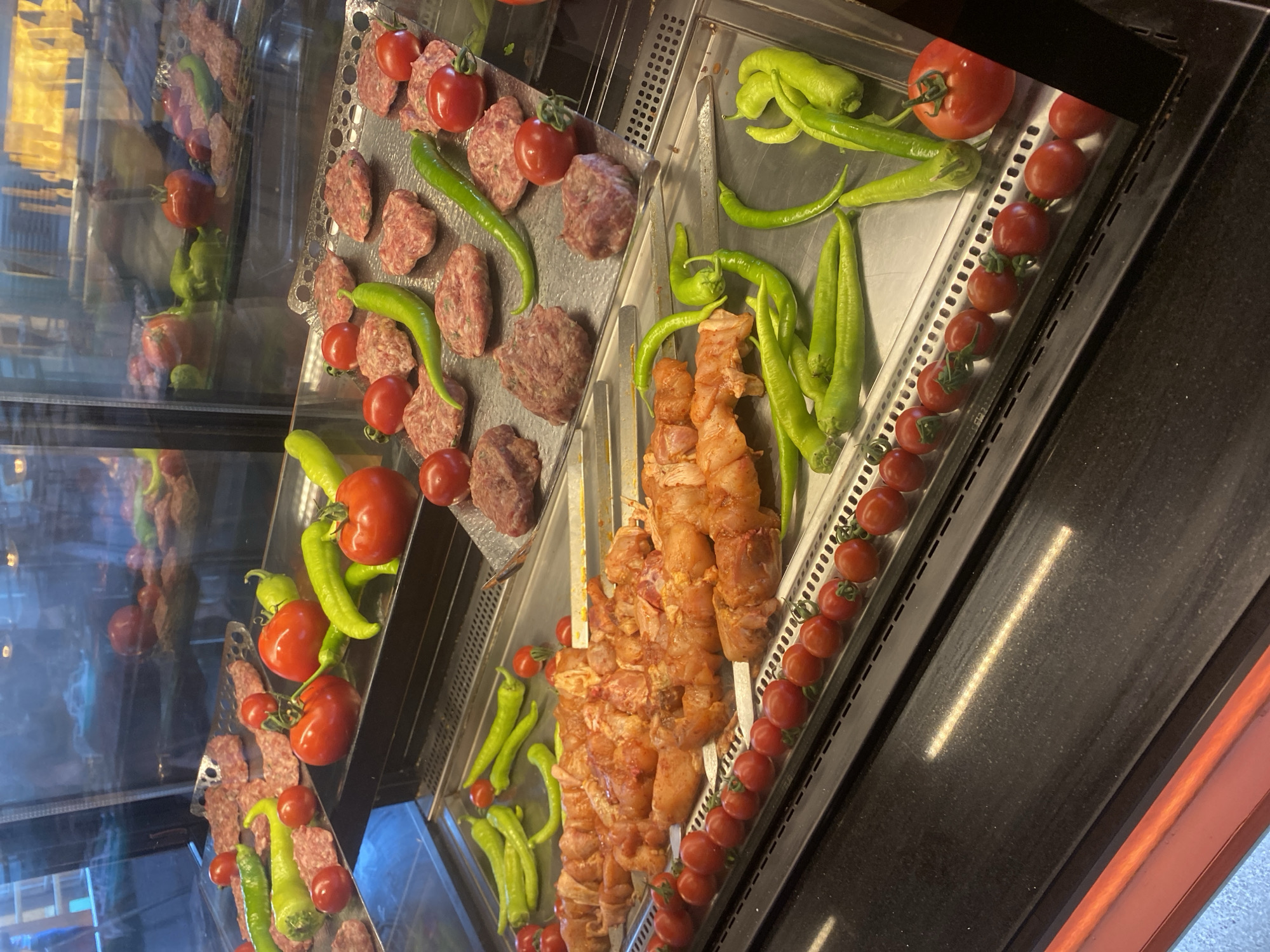 The image shows a display case filled with raw meats and vegetables, likely in a butcher shop or restaurant in Aksu, Turkey. In the foreground, skewers of raw chicken are arranged on a metal tray, surrounded by cherry tomatoes. Behind the chicken, more skewers of meat and peppers are visible, as well as raw ground meat patties and whole tomatoes and peppers. The display case is well-lit, and the overall impression is one of fresh, uncooked ingredients ready for preparation. The reflections in the glass suggest an indoor environment with patrons or staff present, though no individuals are clearly visible.