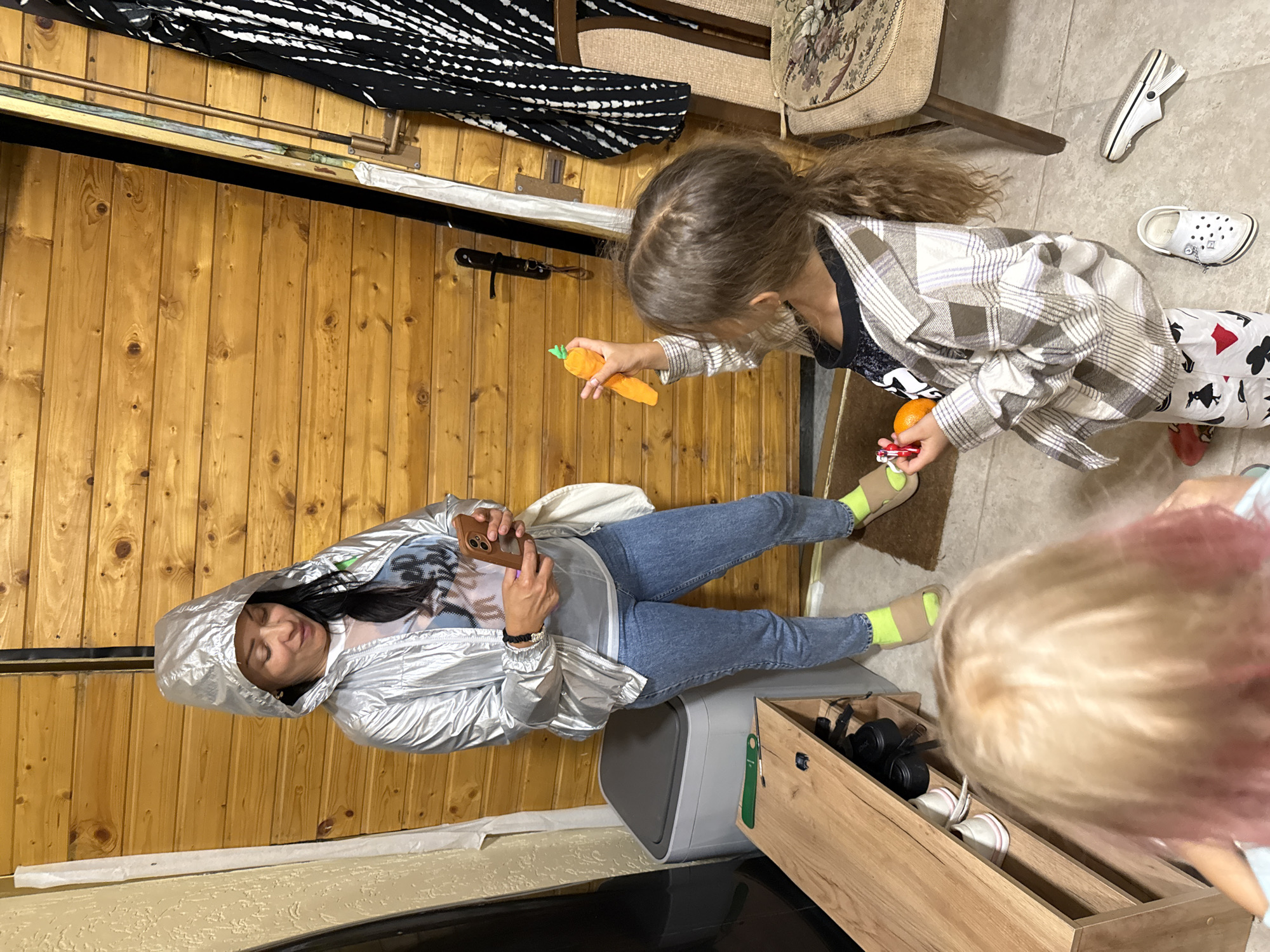 In an indoor scene, a woman wearing a silver hooded jacket, a white graphic t-shirt, blue jeans, and lime green socks stands holding a smartphone. In front of her, a young girl with long brown hair, wearing a plaid shirt and patterned pants, holds a plush orange carrot toy. Another child, with blonde hair that appears to have pink tips, is seen from behind, facing away from the camera. The background consists of a wooden door and a shoe rack with shoes. The environment suggests an informal setting, possibly a home or a storage area. The lighting is moderate, and the overall scene is clear and in focus.