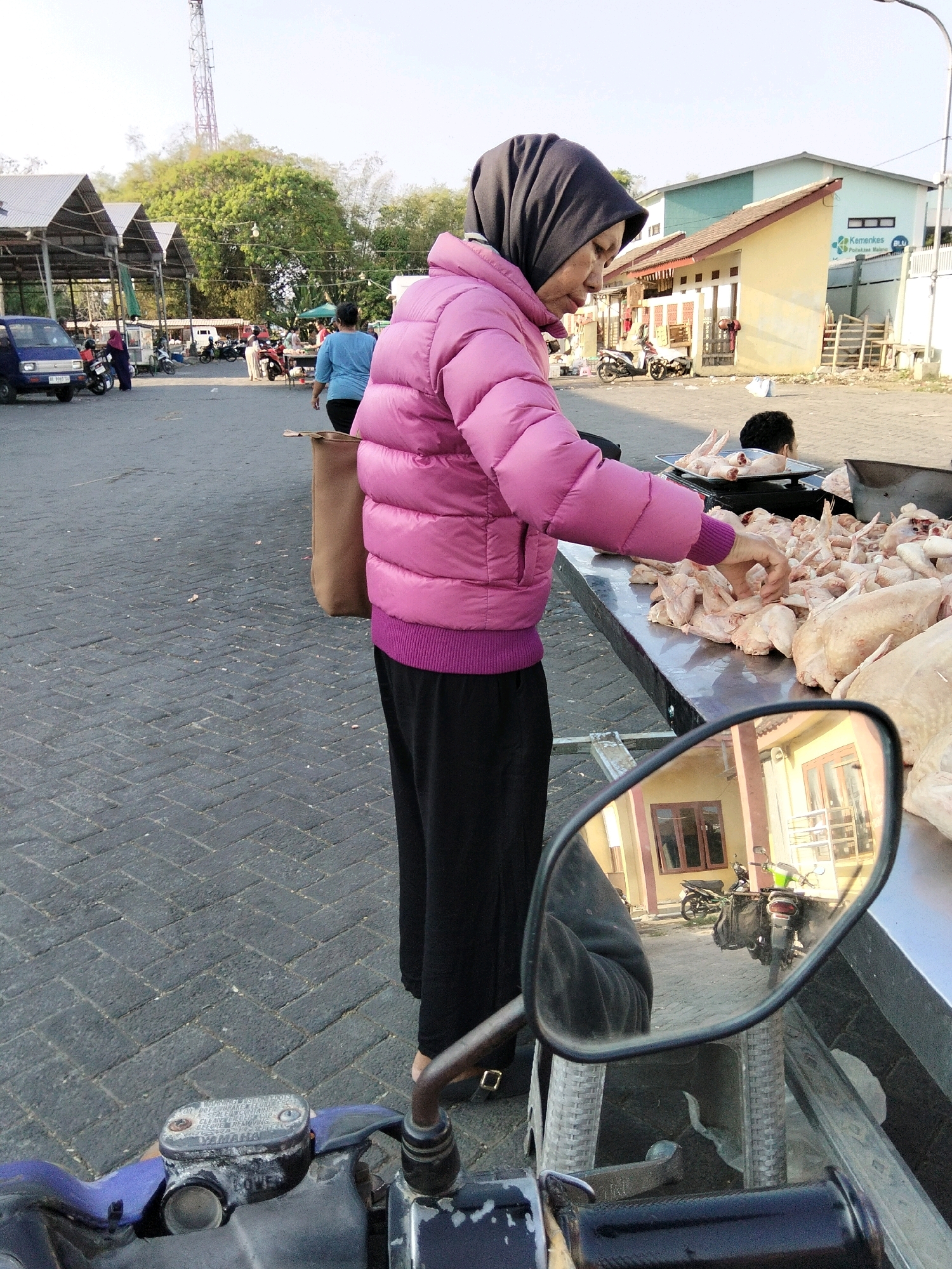 The image depicts an outdoor market scene in Banyudono, Indonesia. In the foreground, a woman wearing a pink puffer jacket and a dark headscarf is seen from the side, reaching towards a large display of raw chicken pieces laid out on a metal table. She is standing next to a motorcycle, the side mirror of which is prominently in the lower portion of the frame, reflecting a building and other motorcycles. In the background, a blue van is parked on a brick-paved surface, and several stalls with pitched roofs are visible, suggesting a bustling marketplace. Other people and motorcycles are scattered throughout the scene, and a tall radio tower is visible in the distance against a bright sky. The overall environment appears to be a public outdoor space, likely a traditional market.