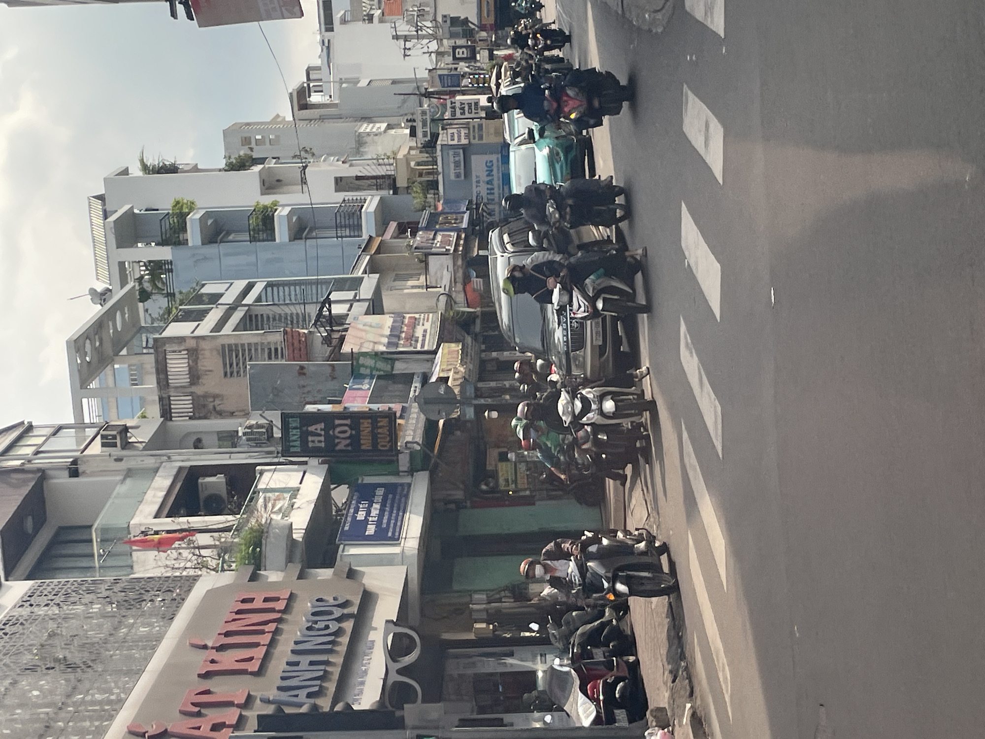 The image depicts a busy street scene in Thủ Đức, Vietnam. A variety of motorcycles and a few cars are navigating the roadway, suggesting moderate traffic. Pedestrians are visible on the sidewalks, some of whom are also on motorcycles. The buildings lining the street are multi-story structures, some with balconies and architectural features characteristic of urban environments in Vietnam. Signage in Vietnamese is present on the buildings, indicating commercial establishments. The weather appears to be overcast, with a bright sky visible. The overall atmosphere suggests a typical day in a bustling Vietnamese city.
