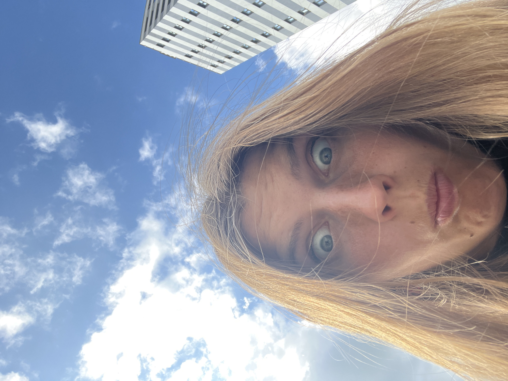 An extreme low-angle, close-up selfie shows a Caucasian woman with blonde hair and wide, surprised eyes looking into the camera. Her mouth is puckered, as if in shock or about to speak. Her hair is windblown, framing her face against a bright blue sky with scattered white clouds. To the right, a tall, modern white building with many windows rises into the sky, characteristic of the architectural style found in cities like Minsk, Belarus. The perspective suggests the photo was taken outdoors, possibly from street level looking upwards.