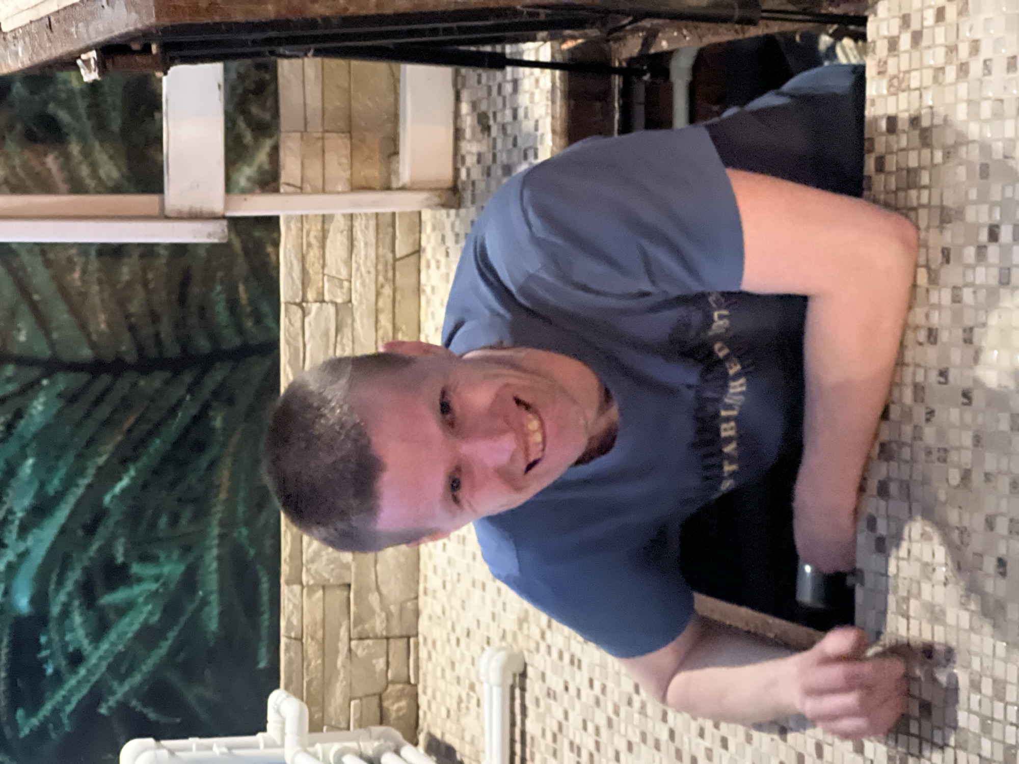A man is positioned within a narrow, tiled space, possibly a maintenance access point in Perm, Russia, as the tiling pattern is characteristic of the region. He is wearing a blue t-shirt and appears to be smiling, looking directly at the camera. A dark object, possibly equipment or clothing, is visible behind him. In the background, there is a wall with light-colored, rectangular tiles and a tank containing synthetic foliage. The surroundings suggest an indoor environment, possibly a utility room or service area connected to a public facility.