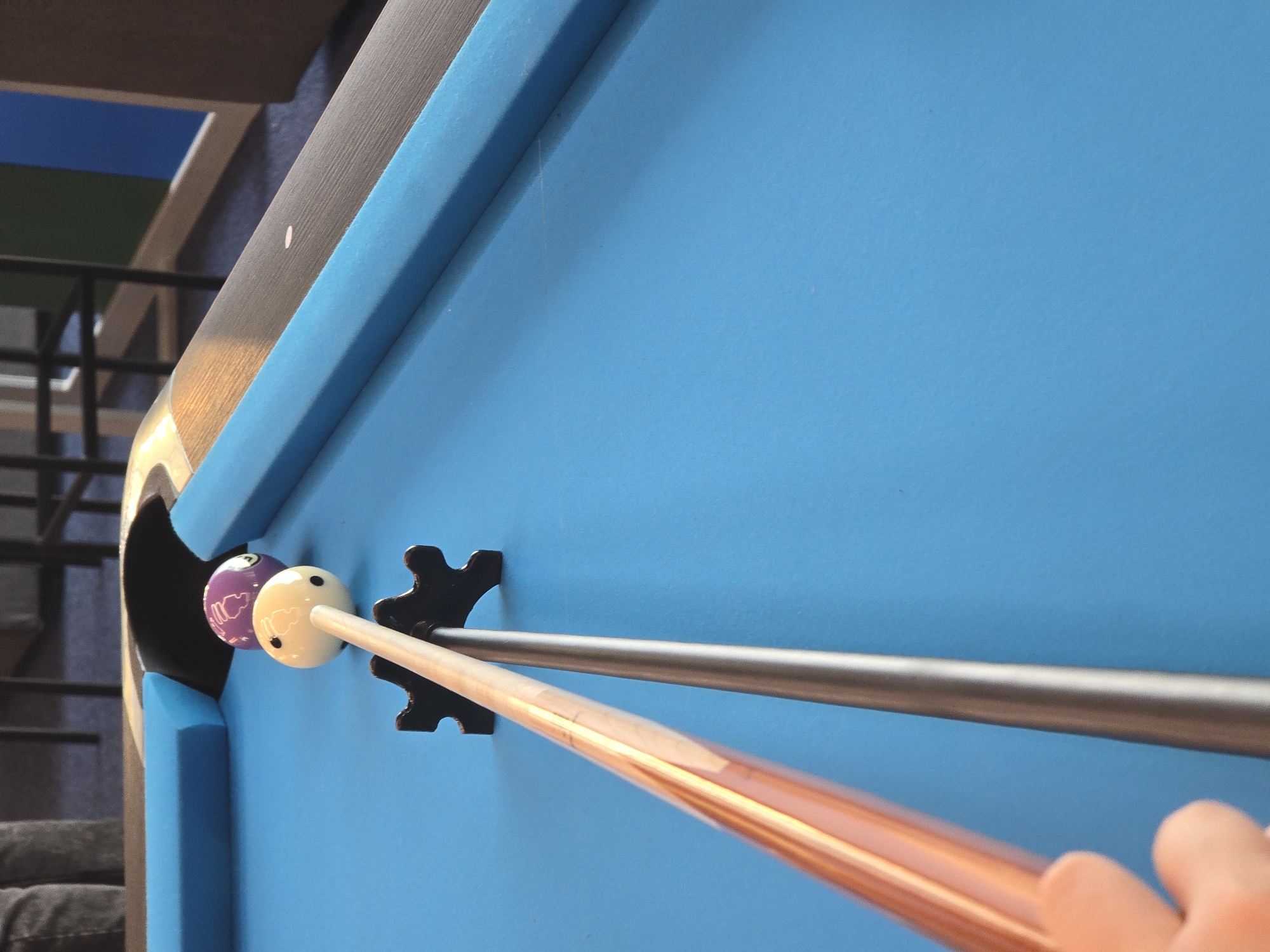 The image depicts a close-up of a pool table. A purple striped billiard ball and a white cue ball are positioned near the corner pocket. Two billiard cues, one metallic gray and one reddish-brown, are positioned through a bridge rest on the table. A person's hand is visible at the edge of the frame, holding one of the cues. The pool table's surface is a bright blue color. The location is unknown and not visibly identifiable as Varna, Bulgaria.