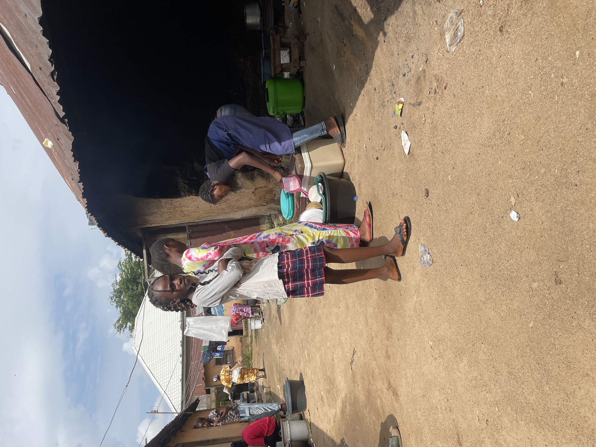 In an outdoor environment in the city of Jos, Nigeria, three young girls stand in a dirt courtyard. The girl on the left, wearing a grey long-sleeved shirt and a plaid skirt, faces the camera with her arms crossed. To her right, a second girl in a brightly colored tie-dye dress stands with her back partially turned. A third girl, wearing a blue apron over jeans and a black t-shirt, is bent over and appears to be washing dishes or preparing food. Several plastic containers, some containing items, are visible near her. The background features rustic buildings with corrugated metal roofs, laundry hanging on a line, and scattered debris on the ground, including pieces of paper and plastic. The overall scene depicts a domestic setting, possibly a communal living area or a market stall.