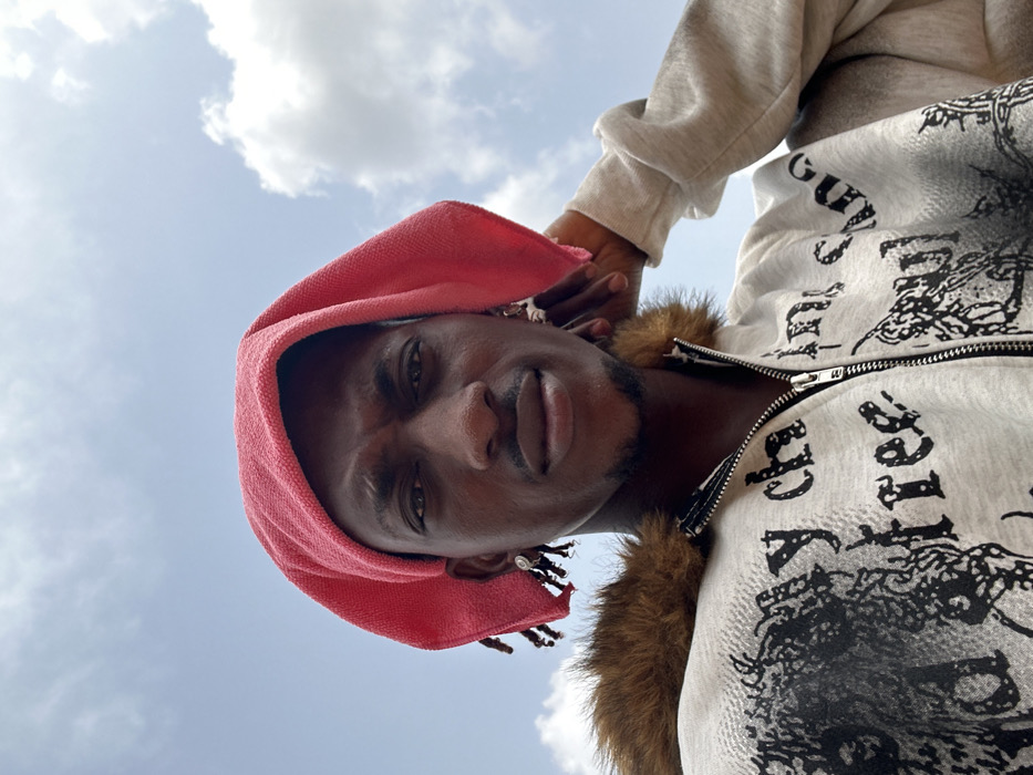The image presents a low-angle, close-up portrait of a single male individual outdoors in Egbe, Nigeria. The subject has dark skin, dark eyes, and a short, dark beard and mustache. A pink, textured cloth or towel is draped over his head, partially obscuring dark dreadlocks or braided hair visible near his ears. Each earlobe features a silver-toned hoop earring with a small attached dangle. He is wearing a light grey hooded sweatshirt or jacket with a front zipper and a prominent brown faux fur trim around the neck/hoodline. The garment is embellished with dark, distressed graphic prints, including fragmented text such as "my ch" and "free". The subject's left arm is raised, with his hand positioned near the back of his head or neck. The background consists of a bright blue sky with dispersed white cumulus clouds.
