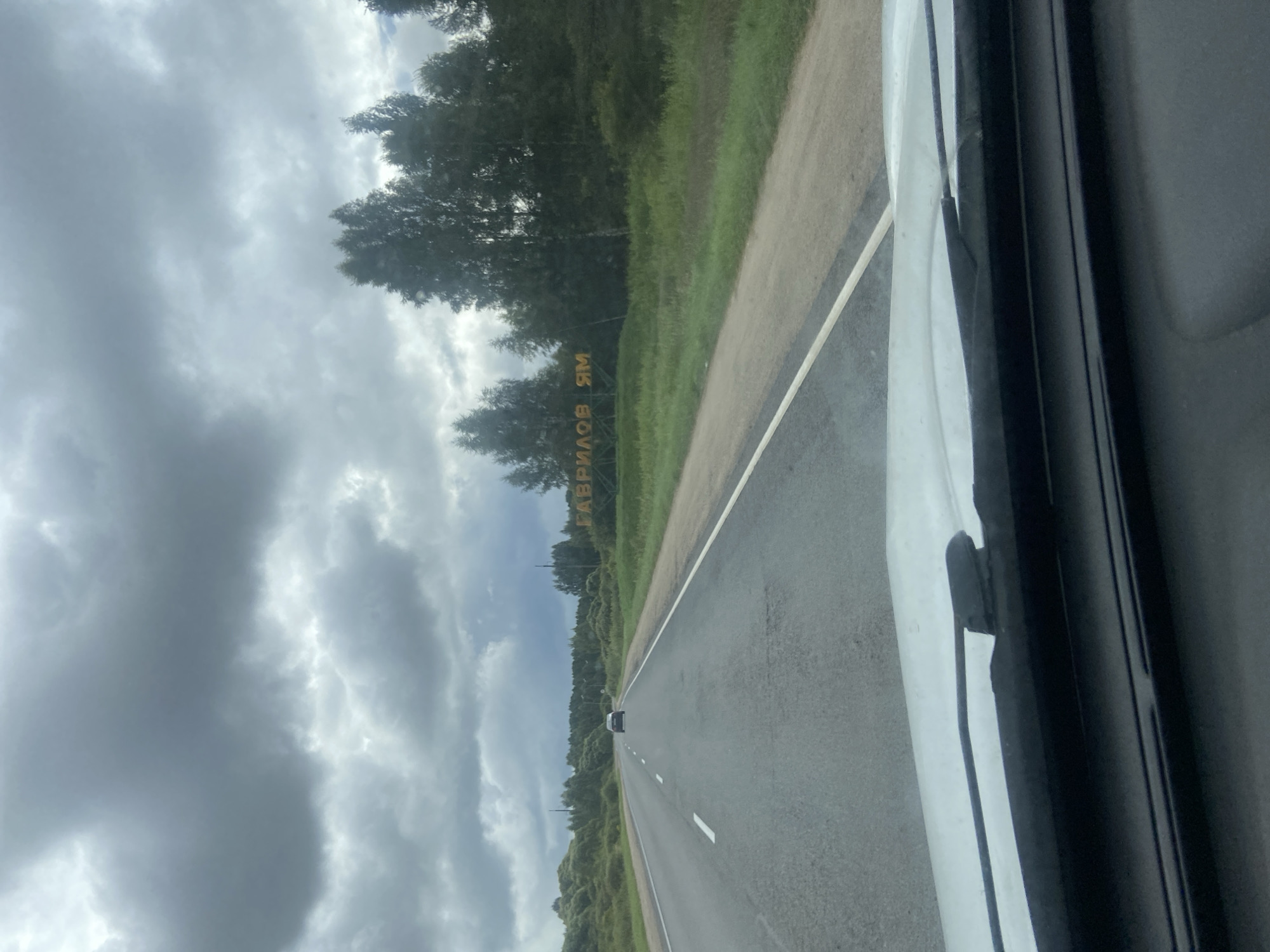 The image shows a view from inside a vehicle looking out at a road in Zayachii Kholm, Russia. A dark-colored car is visible driving ahead on the road. To the right of the road, a large sign with yellow lettering is partially visible, reading "ГАВРИЛОВ ЯМ". This sign is positioned in front of a line of trees and lush green grass. The sky above is overcast with heavy grey clouds. The foreground of the image is dominated by the interior of the vehicle, including the windshield and dashboard.