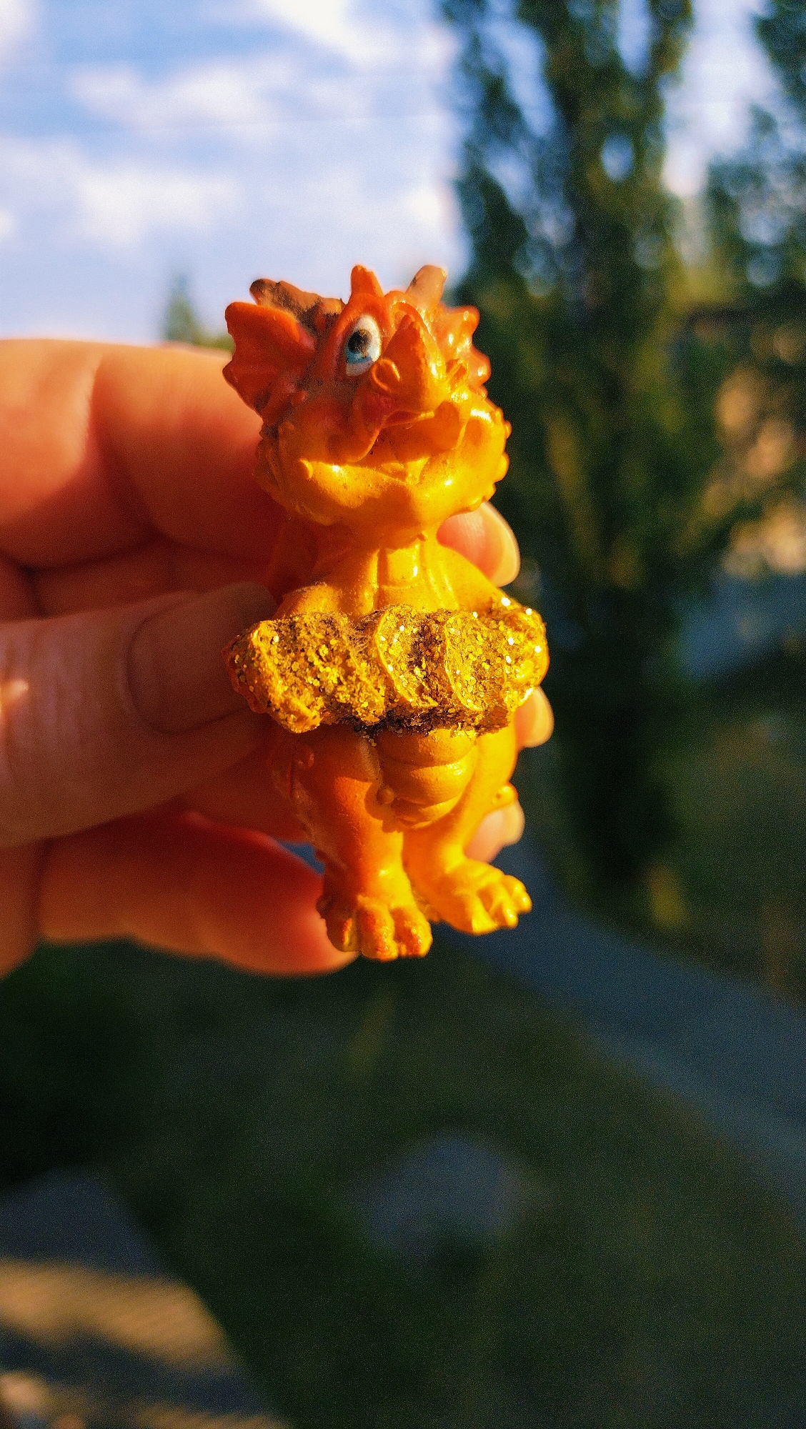A person's hand is holding a small, orange, dinosaur-like figurine. The figurine has a spiky head, a wide smile, and is wearing a glittery, gold belt around its waist. The background is blurred, showing green foliage and a blue sky with clouds, suggesting an outdoor setting. The lighting indicates it may be late afternoon. The image was captured in Horishni Plavni, Ukraine.