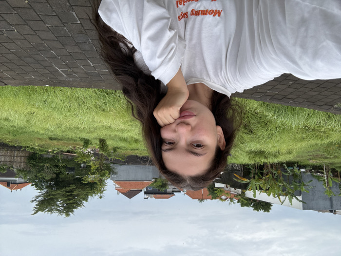 A female subject is depicted in a selfie taken in Dalung, Indonesia, against an outdoor background. The subject, possessing long dark hair, wears a loose-fitting white long-sleeve shirt with the orange text "Mommy Says I'm Special" visible on the front. Her right hand supports her chin, and her lips are slightly puckered. The foreground features a dark grey paved surface composed of rectangular blocks. The mid-ground is dominated by an overgrown field of tall green grass. In the background, a residential area is visible, characterized by multiple houses with orange-tiled roofs, grey and unpainted concrete walls, and abundant tropical vegetation, including several banana trees. The sky is overcast and grey.