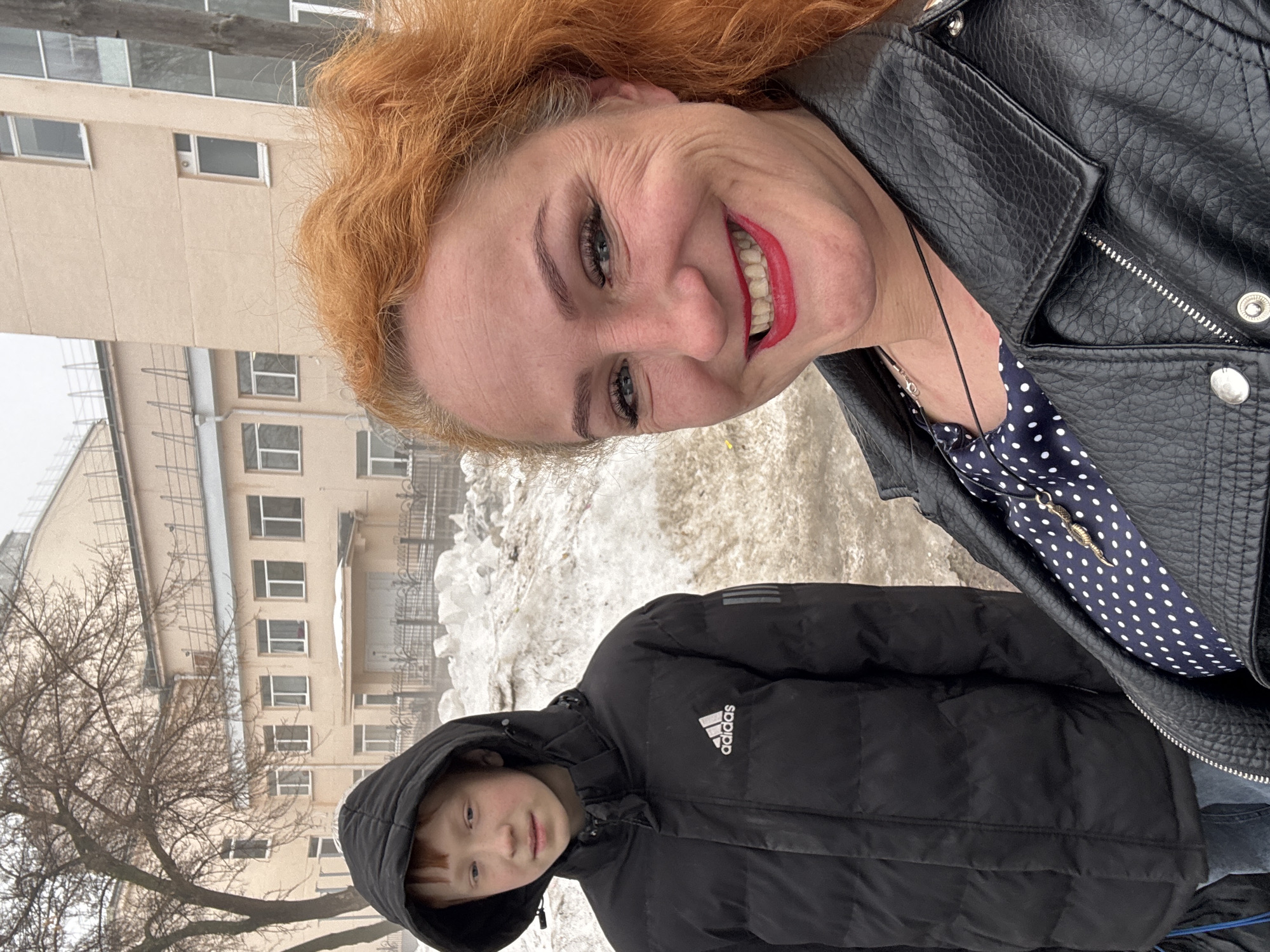 A woman and a child are posing for a selfie outdoors. The woman is red-haired, wearing a black leather jacket over a polka-dot top, and has bright red lipstick. The child is wearing a black Adidas hooded jacket. The background includes a building with multiple windows, some snow, and bare trees. The image was taken in the city of Chaikovsky, Russia.