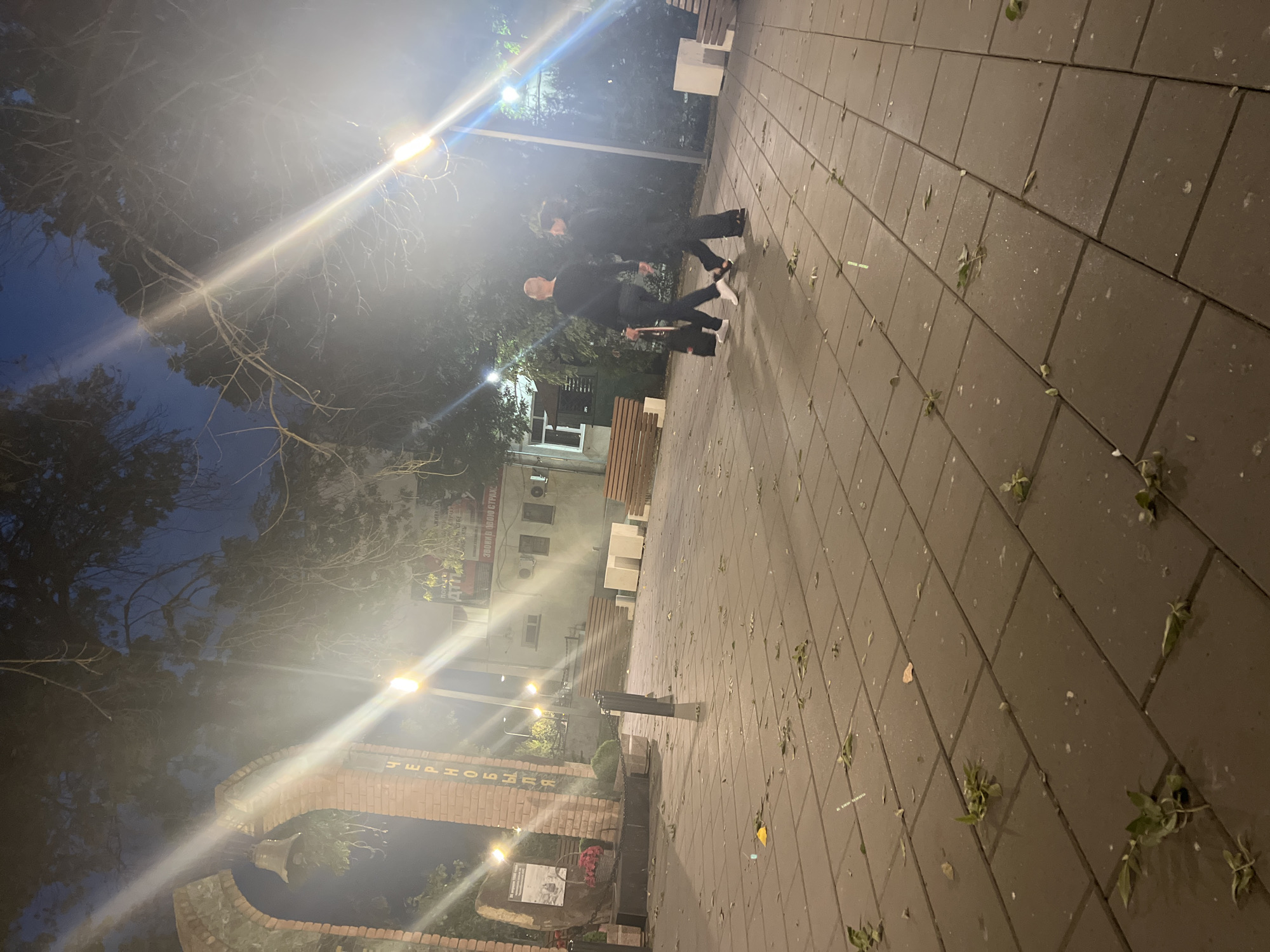 The image depicts an outdoor scene at night, possibly in a public square or park. The ground is paved with rectangular tiles, and scattered leaves are visible on the pavement. In the background, a brick archway with a bell is prominent, with vertical lettering on the brickwork. To the left of the archway, there is a monument or memorial with a stone plaque and a basket of red flowers.
Several streetlights illuminate the area, creating bright flares and reflections in the night sky. Trees with bare branches are visible, their silhouettes against the dimly lit sky. In the mid-ground, two individuals are walking away from the camera. The person on the left, with a bald head, is wearing a dark long-sleeved shirt and dark pants, carrying a black bag. The person on the right is wearing dark clothing.
Further in the background, a building with windows and air conditioning units is present, along with wooden benches and white cubic structures. The overall atmosphere is somewhat hazy, possibly due to mist or light pollution. The location is described as Unknown, Unknown, with no further specific geographical details provided.