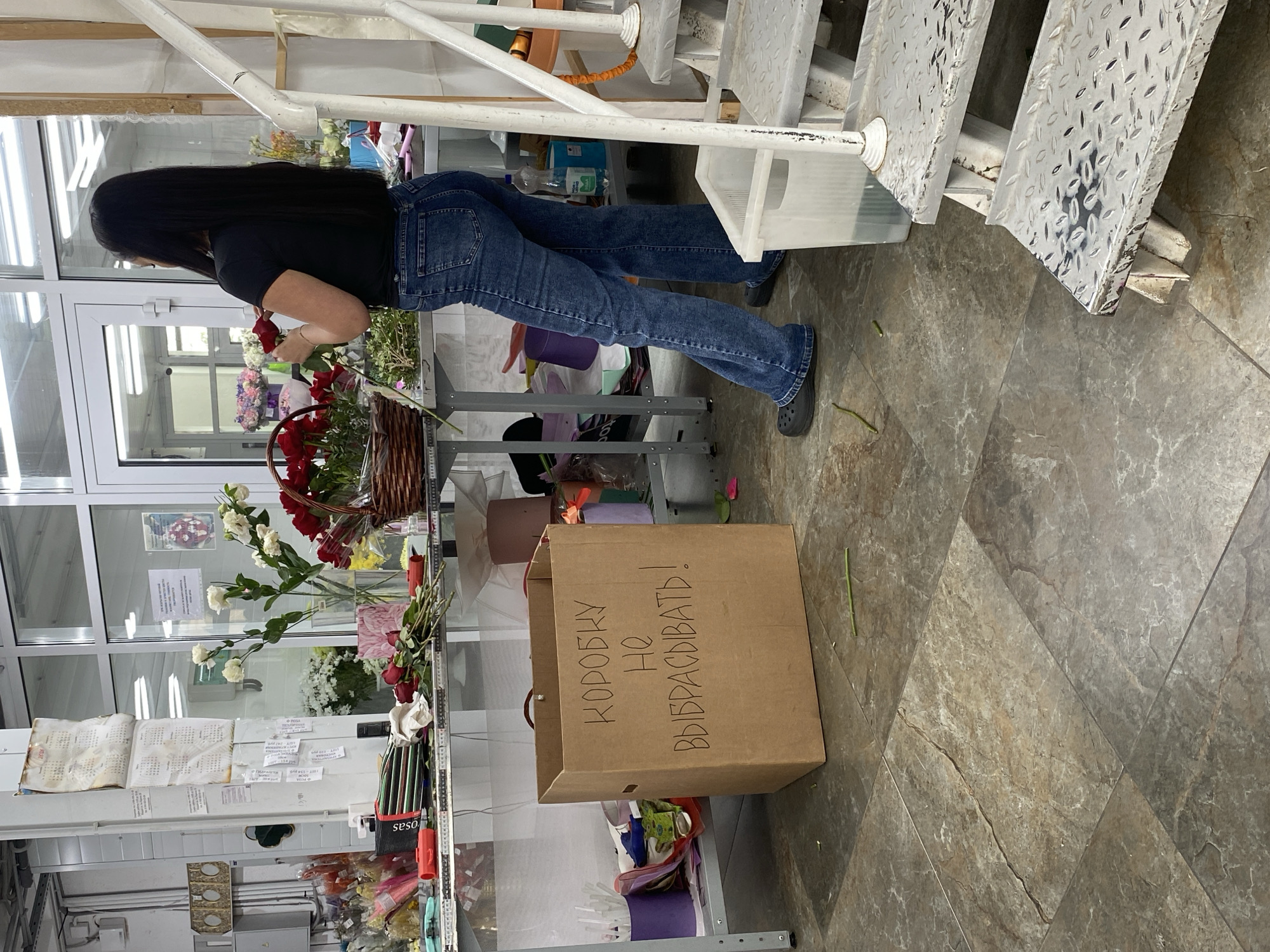 The image shows the interior of a flower shop in Yekaterinburg, Russia. A person with long dark hair, wearing a black shirt and blue jeans, is arranging flowers near a display case filled with various blooms and floral supplies. A large wicker basket filled with red roses is also visible. A cardboard box with the inscription "КОРОБКУ НЕ ВЫБРАСЫВАТЬ!" (Do not throw away the box!) is placed on the floor. The floor is tiled with gray stone-like tiles and the lighting appears to be fluorescent. Metal stairs and white metal railings are also visible.