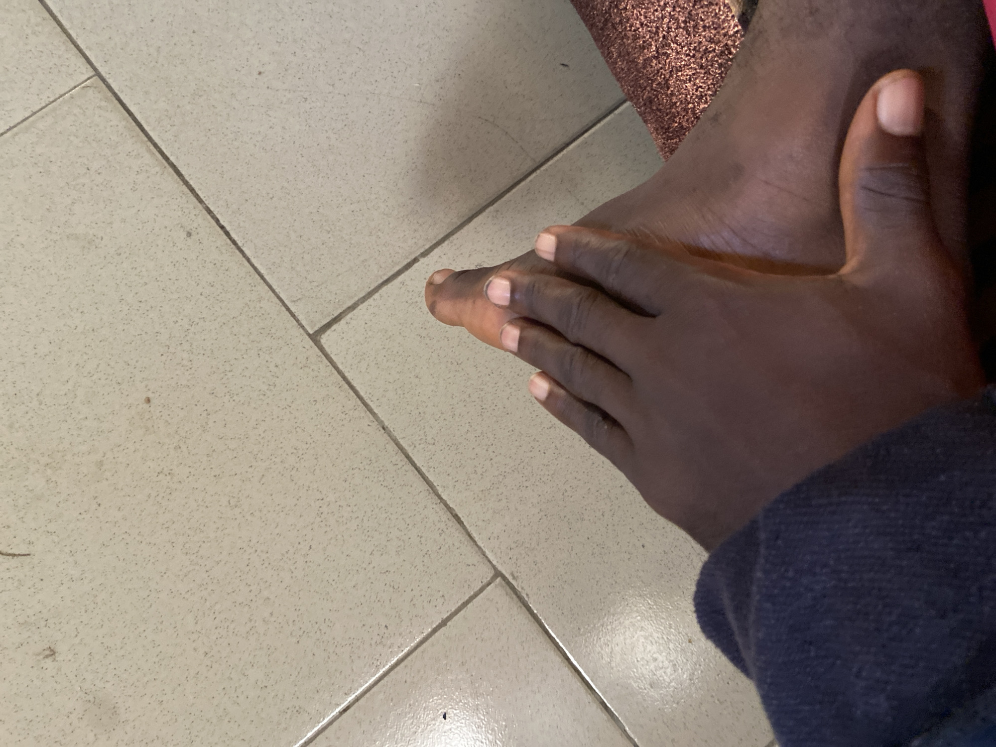 A close-up, overhead shot shows a person's dark-skinned foot, with fingers from the same person's hand gently resting on top of it. The foot is positioned on a tiled floor, with light-colored square tiles arranged in a grid pattern. The tiles have a speckled texture. A dark blue garment is visible in the lower left corner of the image, partially obscuring the person's leg and foot. To the right of the foot, a textured, glittery material, possibly fabric, is visible. The scene appears to be indoors, on a floor likely in Mararaba, Nigeria.