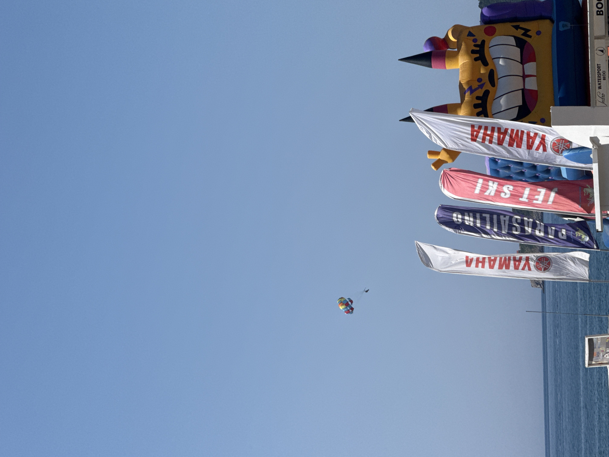 The image captures a bright, sunny day at a beach in Rafailovići, Montenegro. The cerulean sky dominates the upper portion of the frame, with a small, colorful parachute carrying a person visible in the distance. Closer to the foreground, a large, inflatable structure, resembling a cartoon character with a wide, toothy grin and pointy ears, stands prominently. Several vertical flags are positioned in front of the inflatable. One flag is white with red lettering that reads "YAMAHA". Another flag is purple with blue lettering and the word "PARASAILING". A third flag is red with white lettering that says "JET SKI". The sea, a deep blue, stretches across the lower part of the image, meeting the horizon. The beach area appears to be a hub for water sports, indicated by the flags and the inflatable attraction.