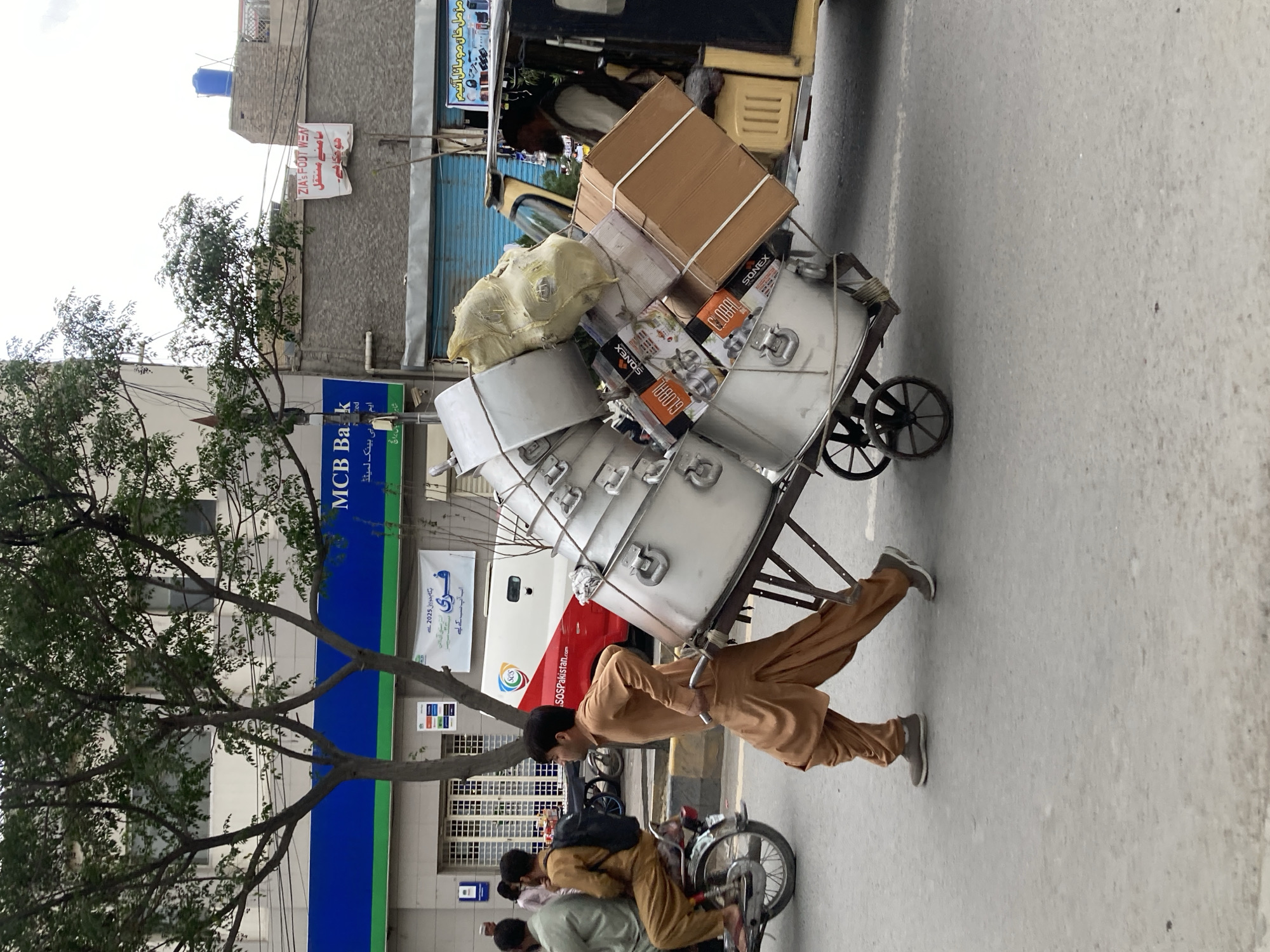In a bustling street scene in Quetta, Pakistan, a man in a light brown shalwar kameez pushes a heavily laden handcart. The cart is piled high with several large, cylindrical metal containers, possibly cooking pots or storage vessels, along with numerous cardboard boxes and a light-colored sack. Some of the boxes are visibly branded "GLOBAL" and "SONEX."
The man is actively pushing the cart forward, indicating a task of transportation or delivery. In the foreground, other men are seen on a motorcycle, suggesting regular street activity.
The setting is an urban street flanked by buildings. A prominent blue sign for "MCB Bank Limited" (ایم سی بی بینک لمیٹڈ) is visible. Other signs, some in Urdu, include "ZIA'S FOOT WEAR" with text about relocation (سامنے منتقل) and a sign for "مزمل خان موبائل آنیم" (Muzammil Khan Mobile On Line/Antiques). A white van with "SOSPakistan.com" and the SOS logo is also visible. The architecture suggests a mix of commercial and residential structures. Trees with green foliage line the street. The light appears diffused, possibly indicating an overcast day.