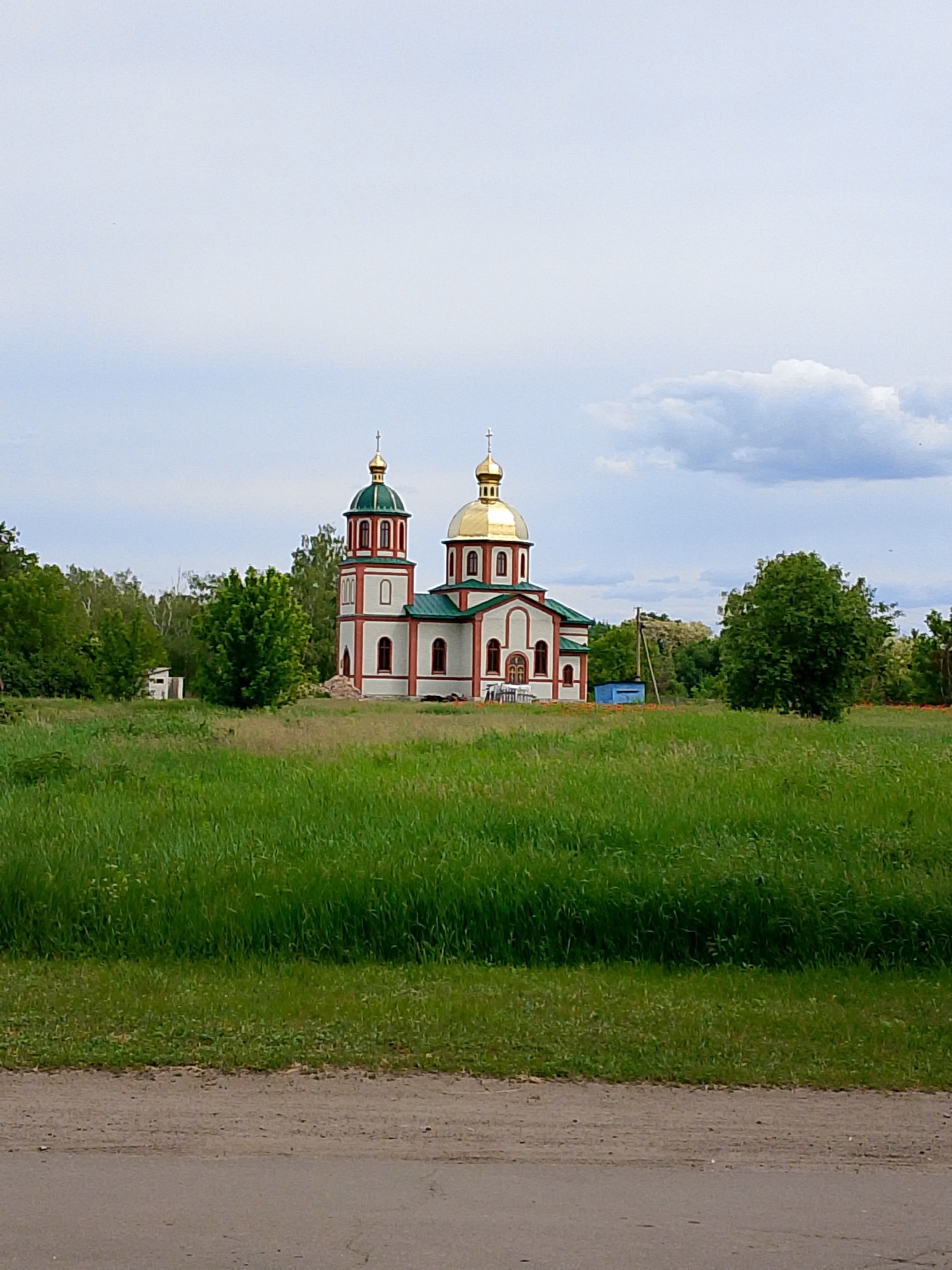 The image presents a serene outdoor scene featuring an Eastern Orthodox church set against a backdrop of lush greenery and a partly cloudy sky. The church is the central subject, characterized by its white walls with red decorative trim and green roofs. It boasts two prominent onion domes, one larger and golden, and another smaller and green, both surmounted by golden crosses.
The church is surrounded by vast expanses of tall, vibrant green grass and numerous trees, suggesting a rural or semi-rural setting. To the left of the church, a small, white, shack-like building is partially visible among the trees. On the right side, behind the church, a small blue box-like structure can be seen. In the immediate foreground, a dirt road or path stretches across the bottom of the frame.
The weather appears to be overcast or partly cloudy, with a bright, diffused light indicating daytime, likely late morning or early afternoon. There are no people visible in the image, and no specific activity or event is taking place, contributing to a calm and tranquil atmosphere. The architectural style of the church, particularly the onion domes, is indicative of Eastern European or Russian religious architecture. No text is visible within the image.