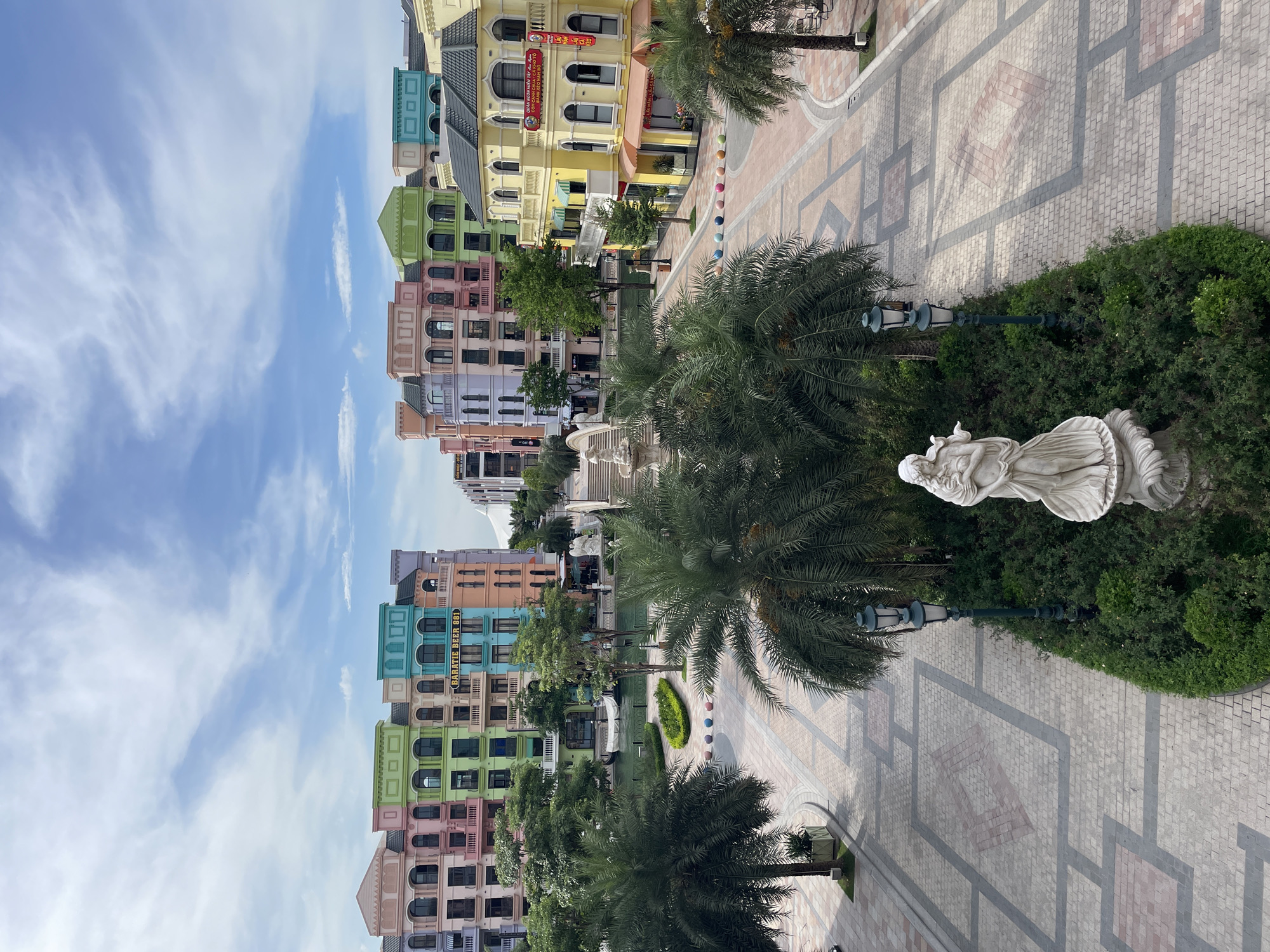 This image depicts a vibrant, European-style street scene in Nghĩa Trụ Commune, Vietnam, likely within the Sun Signature Entertainment complex or a similar resort. The architecture features rows of colorful buildings with pastel facades in shades of pink, green, blue, and yellow, designed with ornate window frames and balconies. Palm trees are interspersed throughout the paved pedestrian area, which is laid with patterned stone tiles. In the foreground, a white marble statue of a draped female figure stands on a pedestal amidst lush green bushes. Two dark green lampposts are positioned symmetrically on either side of the statue. The background reveals a canal with a small white boat, and further down the street, more brightly colored buildings and a fountain are visible. The sky is partly cloudy with a bright blue expanse. No people are visible in the immediate foreground or engaging in any action. The scene conveys a clean, well-maintained, and picturesque tourist destination.