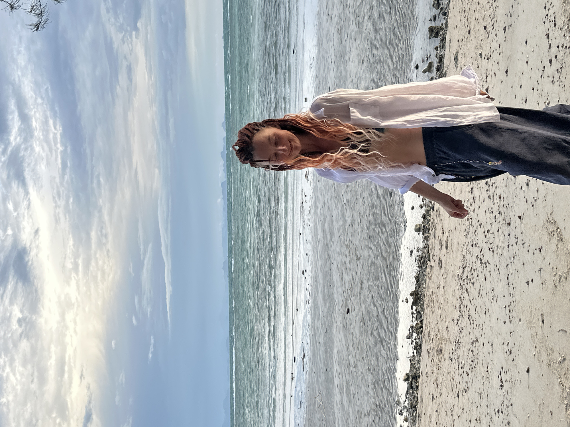 A person with braided hair, which transitions from brown to pink, walks on a sandy beach. They are wearing a white open shirt over a dark bikini top and dark loose-fitting pants. The background shows the ocean with waves and a hazy sky with clouds. Distant landmasses are visible on the horizon. The location is the beach area of Ko Samui District, Thailand.