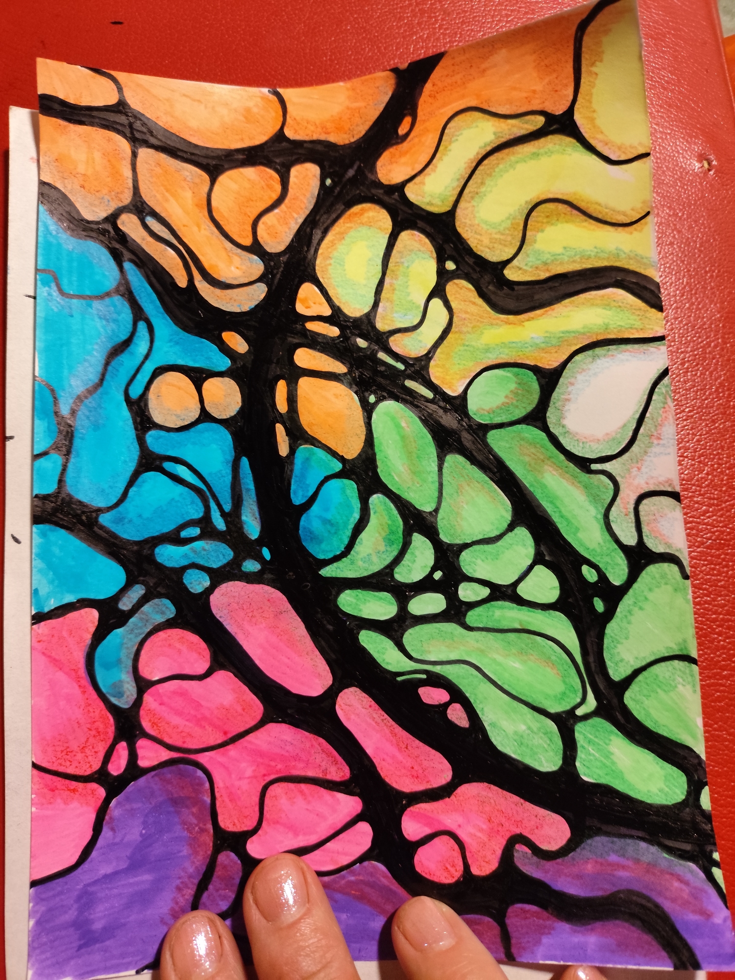 A close-up, top-down, eye-level shot shows a colorful abstract drawing with black lines forming cells filled with various vibrant colors such as orange, yellow, green, blue, pink, and purple. The drawing is on white paper and is placed on a textured red surface, possibly a binder or a table. The image is partially cut off on the right side, revealing the red surface. The bottom of the image shows two fingers with manicured nails, with the fingertips resting on the drawing and the red surface respectively. The drawing appears to be a neurographic art piece, characterized by its organic, flowing shapes. There is no indication of the location being Krasnodar, Russia.