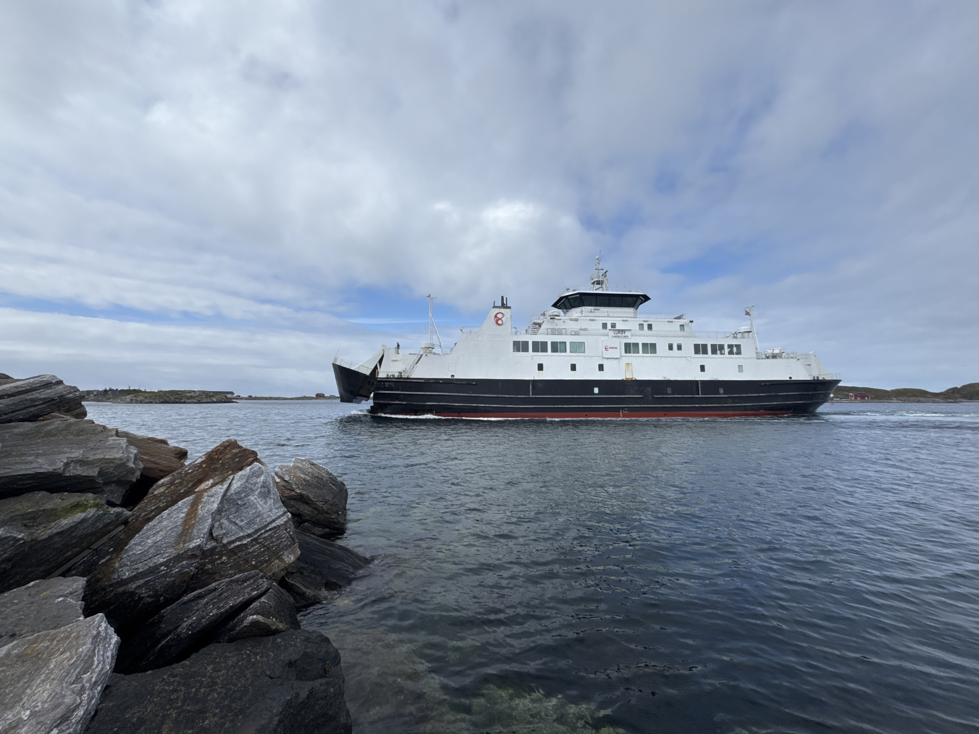 A ferry, identified by the name "LUROEY" and the number "8" on its funnel, sails on choppy blue water towards the right of the frame. The ferry is white with a black hull and a red waterline. The sky above is overcast with textured white clouds. In the foreground, a pile of large, grey, and brown rocks juts into the water from the left side of the image. In the background, a small, treeless island with a few scattered red buildings is visible. The scene appears to be a coastal area in Lovund, Norway. There are no people visible in the frame.