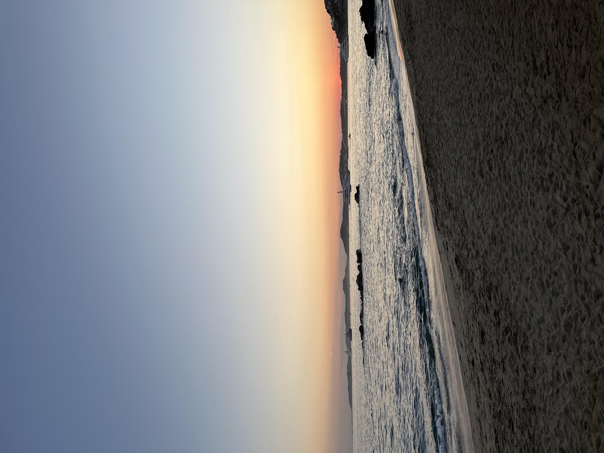 This image depicts a serene beach scene at sunset in Ornos, Greece. The foreground is dominated by a sandy beach, with subtle footprints and the gentle lapping of waves. The sea stretches towards the horizon, reflecting the soft hues of the setting sun. In the distance, a line of hills forms the coastline, dotted with buildings. Several yachts are anchored offshore, adding a touch of luxury to the tranquil landscape. The sky transitions from a pale blue at the top to a warm gradient of orange and pink near the horizon, indicating the end of the day. The overall impression is one of peace and natural beauty.