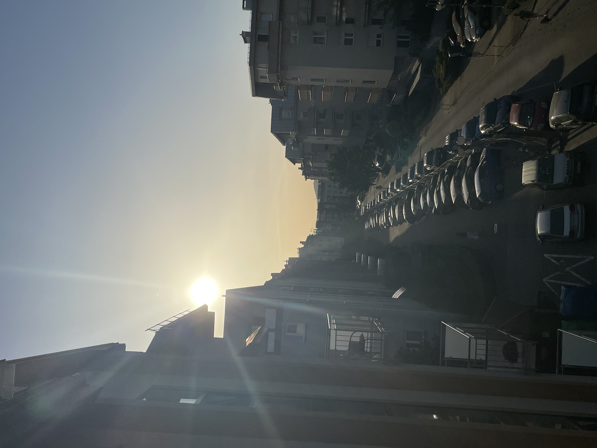The image shows a street in a city lined with buildings. Cars are parked on both sides of the street. The sun is low in the sky, creating a hazy or foggy atmosphere. The buildings are tall and have multiple windows. The city appears to be residential. The exact location is unknown.