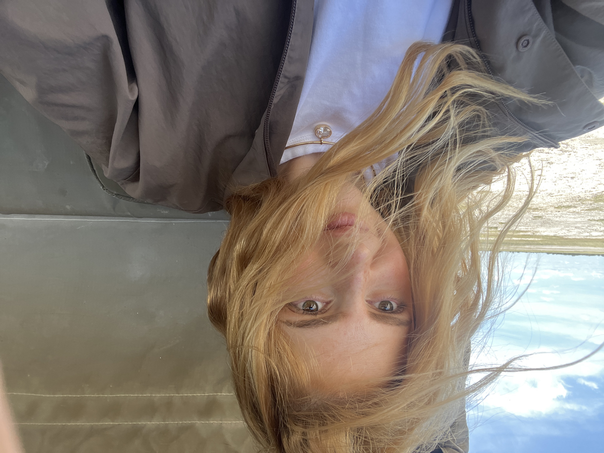 A woman with blonde hair is pictured from a lower angle, so the bottom of her face is closest to the camera and the top of her head is further away. Her hair is tousled, with strands partially obscuring her face. She has light skin and hazel eyes. She wears a white t-shirt and a fine gold chain necklace with a small pendant. Over the t-shirt is a brown, buttoned jacket. The background shows a blurred landscape of low vegetation and sky. The picture appears to be taken outdoors in the vicinity of Gucta, Botswana.