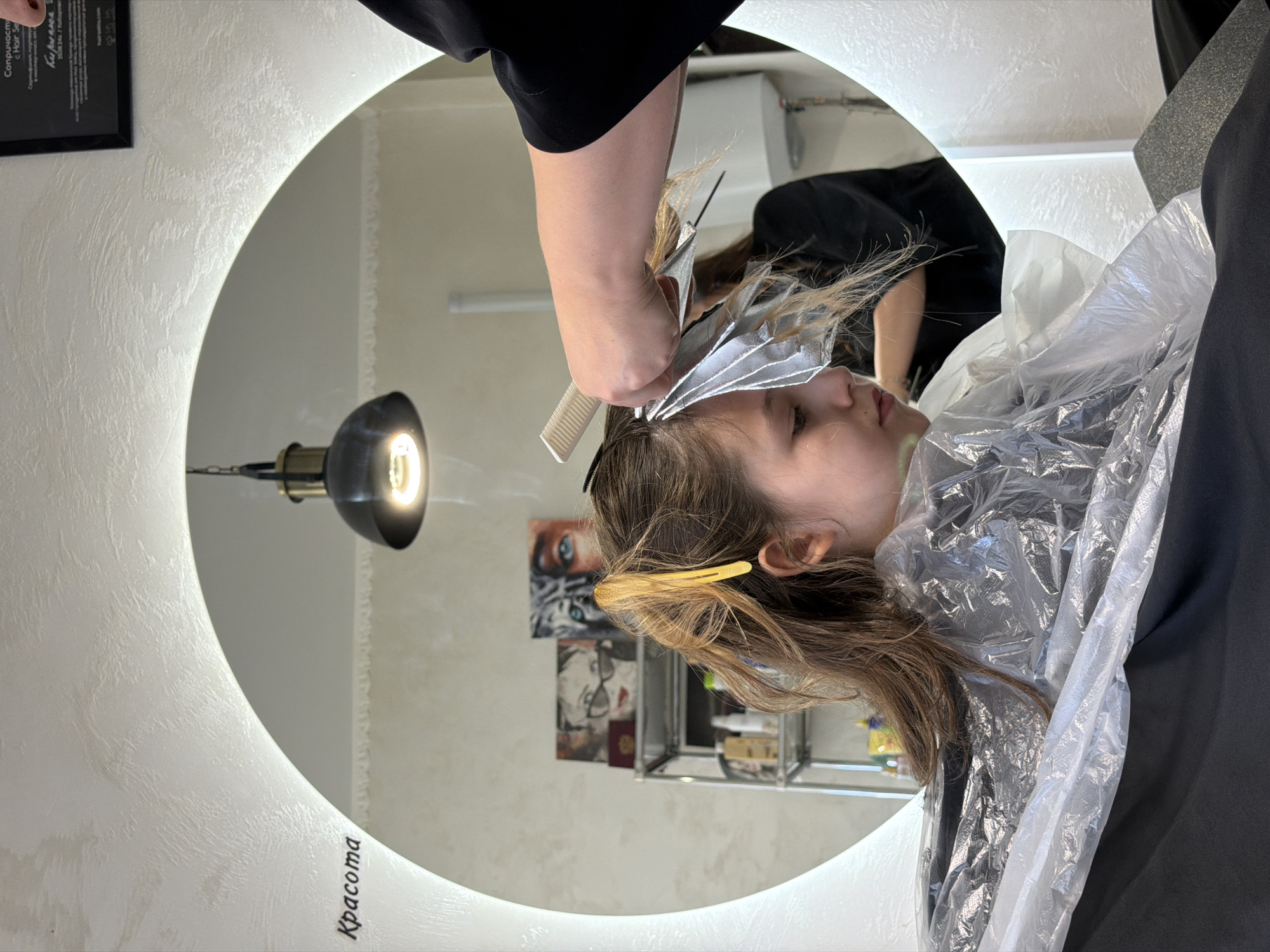 A woman is getting her hair highlighted. The photograph is framed by a round mirror with built-in lighting. She has been draped with a plastic cape. A hairdresser is holding foils and combing her hair. The photo seems to be taken in a salon in Birobidzhan, Russia, due to the cyrillic text visible in the image. The salon has beauty products and paintings on the wall.