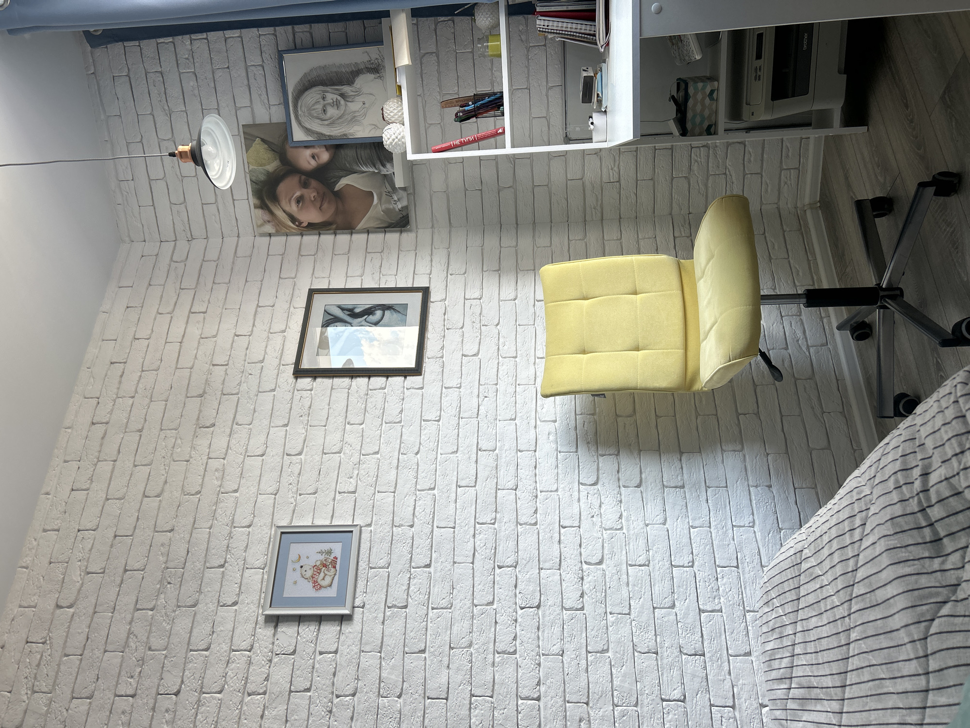 The image shows a corner of a room, likely a bedroom or study, with a white brick-patterned wall. On the wall, there are several framed pictures, including a small cross-stitch of a bear, a drawing of a face, and a photograph of a woman and child. A yellow office chair is placed in the foreground, facing a white desk. The desk has a printer underneath and a shelf above it, stocked with books and stationery. A blue curtain is visible on the right side of the image. The floor is a dark wood laminate. The lighting appears to be natural, coming from a window that is not visible in the frame. There are no people visible in the image. The overall impression is of a tidy and personalized living space. The location in Orenburg, Russia is not discernible from the visual information provided in the image.