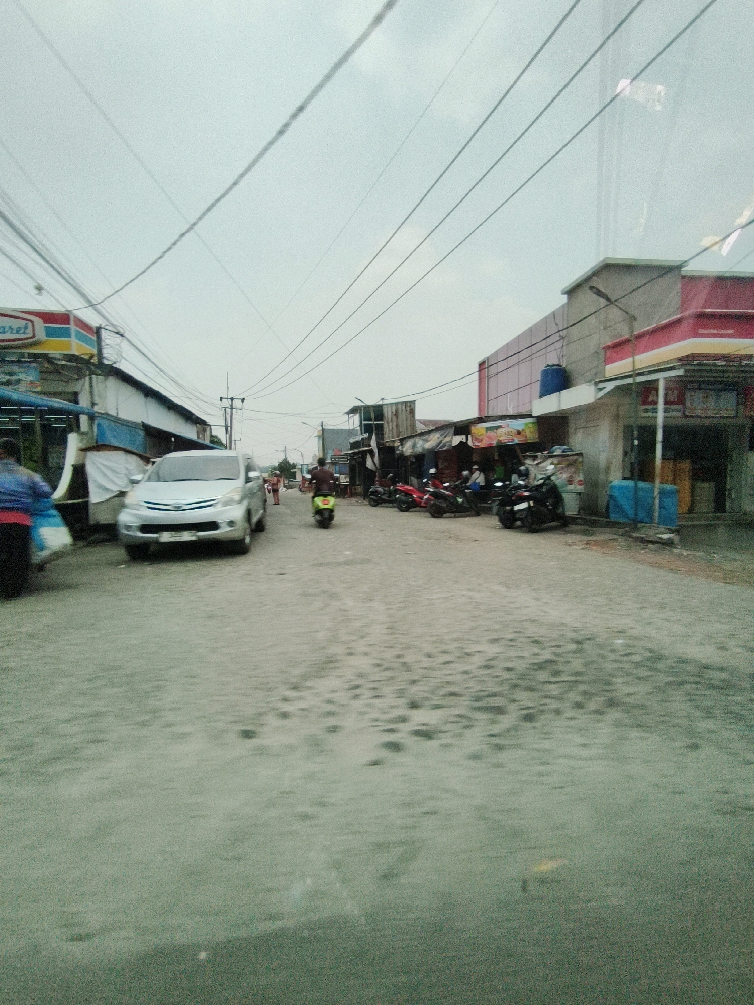 The image depicts a street scene in Sukaraja, Indonesia, likely a commercial area with several shops and parked motorcycles. In the foreground, a light-colored car is driving down the street, which appears to be paved with cobblestones or rough asphalt and has some puddles or wet patches. On the left side of the street, there is a Minimart branded "ALFAMART" with its red and yellow signage. Further down the street on the left, there are more stalls and shops. On the right side of the street, there are several parked motorcycles, and buildings with signage, one of which appears to be another retail establishment with a red and white facade. Power lines are visible crisscrossing the sky. A person wearing a blue shirt and red pants is walking away from the camera on the left side, carrying two white bags. Another person is seen riding a green motorcycle further down the street on the right. The sky is overcast, suggesting a cloudy day.