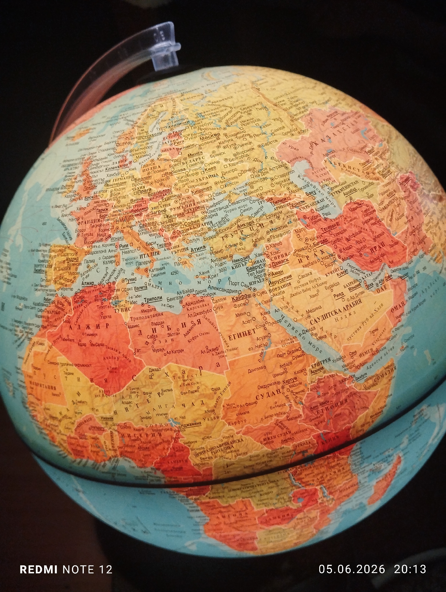 A photograph captures a close-up, angled view of a physical globe, primarily showcasing portions of Europe, Africa, and Asia. The globe is rendered in various colors, distinguishing different countries, with cities and geographical features labeled in Cyrillic script.
In the upper left quadrant, parts of Western Europe are visible, including the United Kingdom, France, Spain, Portugal, Germany, Italy, and countries in Scandinavia and Eastern Europe such as Poland, Ukraine, and Russia. Key cities like London, Paris, Rome, Berlin, Moscow, and Kyiv are marked with circular icons and text.
The central and lower portions of the globe feature North, West, East, and Central Africa, alongside the Middle East and parts of Central Asia. Prominent African countries include Algeria, Tunisia, Libya, Egypt, Sudan, Niger, Nigeria, Ethiopia, and Kenya, with their respective capitals such as Algiers, Tripoli, Cairo, Khartoum, and Addis Ababa. The Middle East displays Saudi Arabia, Iran, Iraq, Turkey, Syria, and Israel, with cities like Riyadh, Tehran, Baghdad, and Jerusalem identified. Central Asian nations like Kazakhstan, Uzbekistan, Turkmenistan, and Afghanistan are also visible.
Bodies of water such as the Mediterranean Sea, Black Sea, Caspian Sea, Red Sea, and Persian Gulf are depicted in blue/teal. A clear, translucent plastic arc, likely part of the globe's stand, extends across the top of the image. The background is uniformly dark, suggesting the globe is illuminated from the front or above, casting soft light across its surface.
There are no individuals present in the scene. The globe is the sole subject. The image includes a digital watermark in the lower left corner reading "REDMI NOTE 12" and in the lower right corner, "05.06.2026 20:13," indicating the device used and the date and time the photograph was captured. The exact location within a city is unknown.