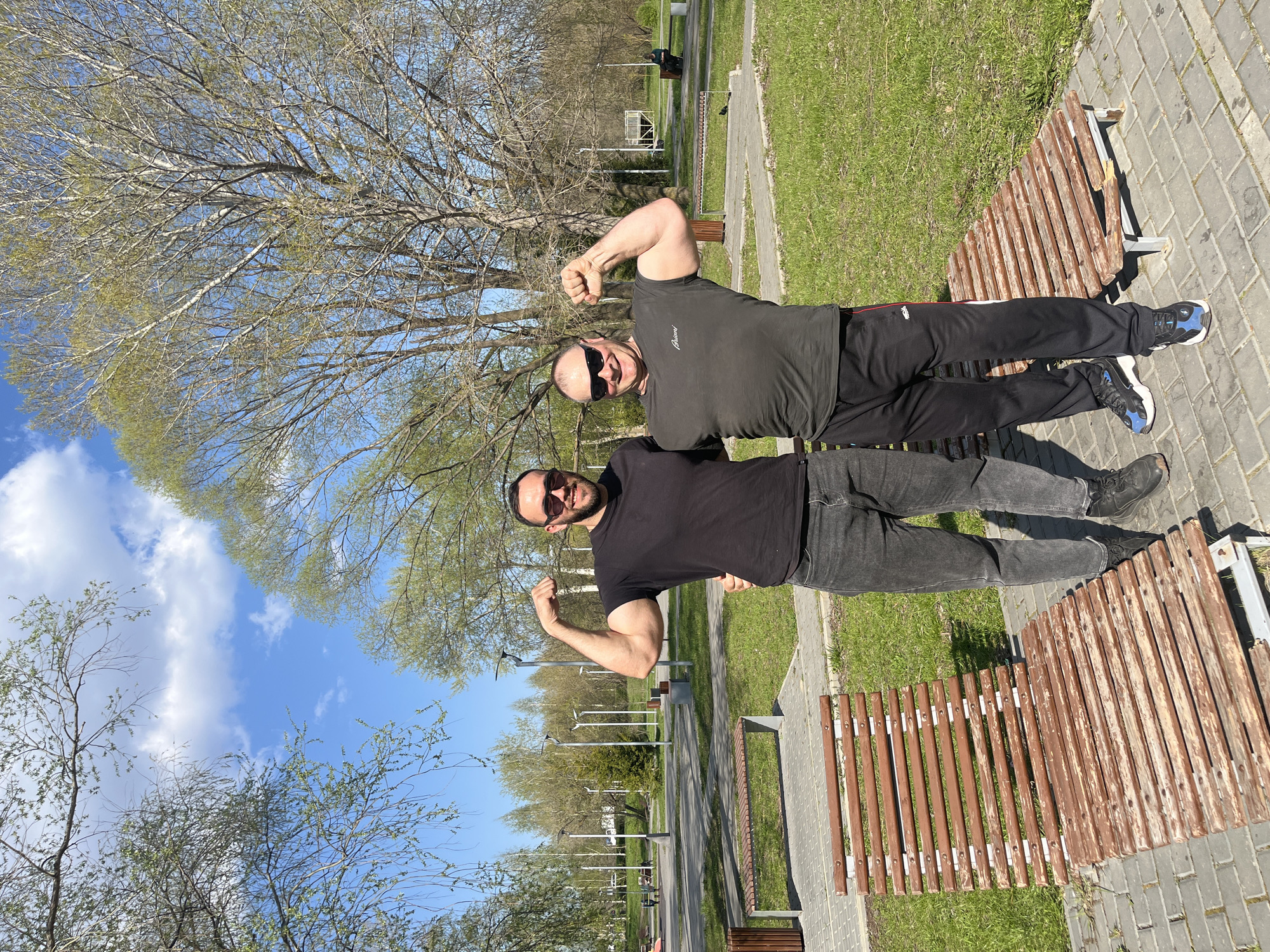 Two adult males are posing on a park bench. The man closer to the camera is wearing a black t-shirt, grey jeans, and black shoes. He is flexing his right bicep and smiling. The other man is wearing a dark gray shirt, black athletic pants, and black and blue athletic shoes. He is also flexing and wearing sunglasses.
They are sitting on a wooden slat park bench with a metal frame. The ground is paved with grey bricks. There is grass and trees in the background. The sky is blue with white clouds. The image appears to have been taken in a park setting in Ulyanovsk, Russia.