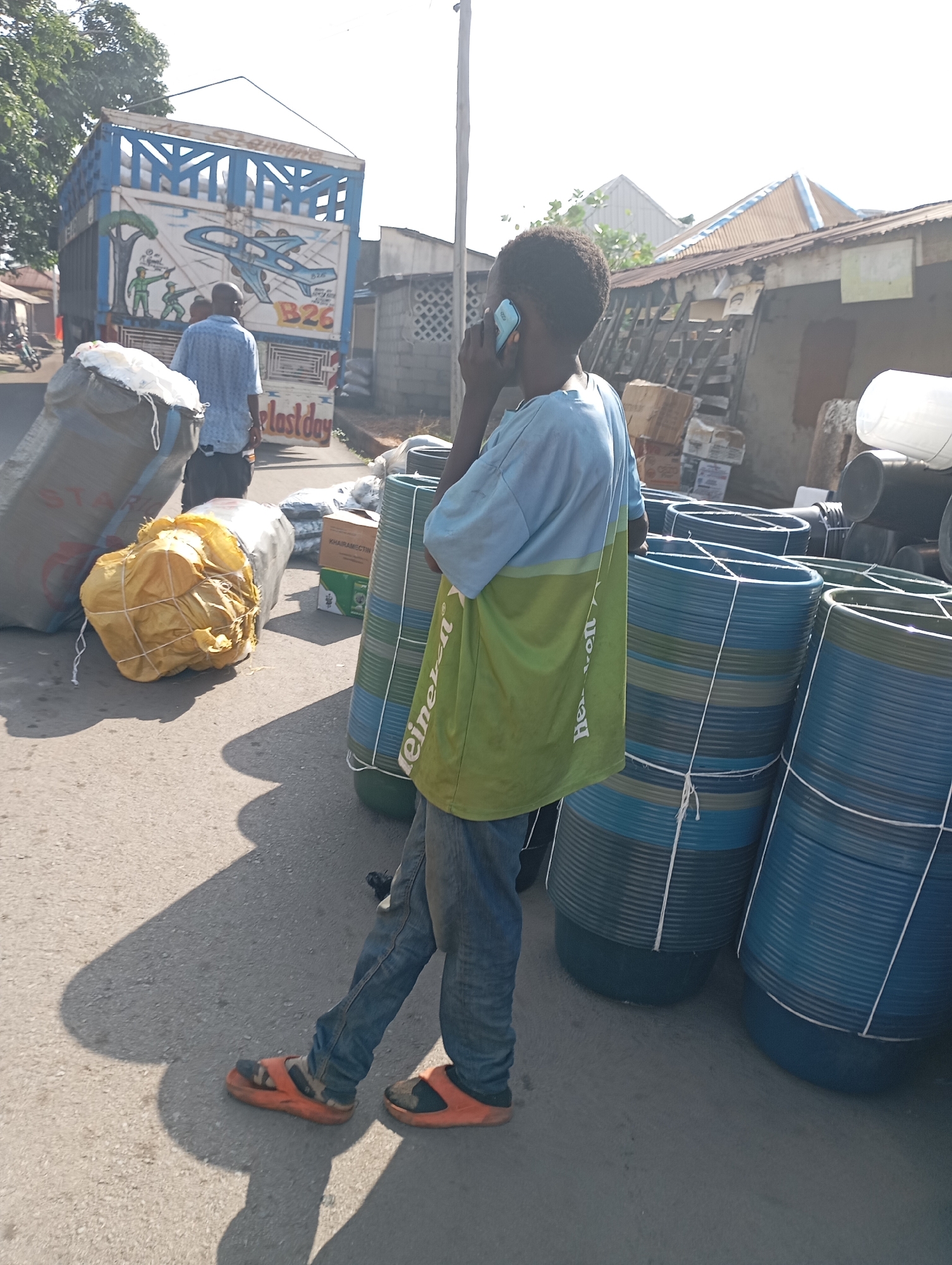 In Shendam, Nigeria, a young boy, observed from behind, stands near a roadside collection of large blue plastic containers. He is dressed in jeans and a blue and green t-shirt, talking on a light blue mobile phone held to his right ear. The boy wears orange sandals with black accents. Adjacent to him are stacks of the plastic containers, some tied together with white rope, and a large yellow bundle wrapped in twine. In the background, a truck with a blue and white painted body featuring an airplane graphic is parked. A man in a blue patterned shirt stands near the truck. The scene appears to be an outdoor market or storage area, with buildings and vegetation visible in the distance under a bright sky.