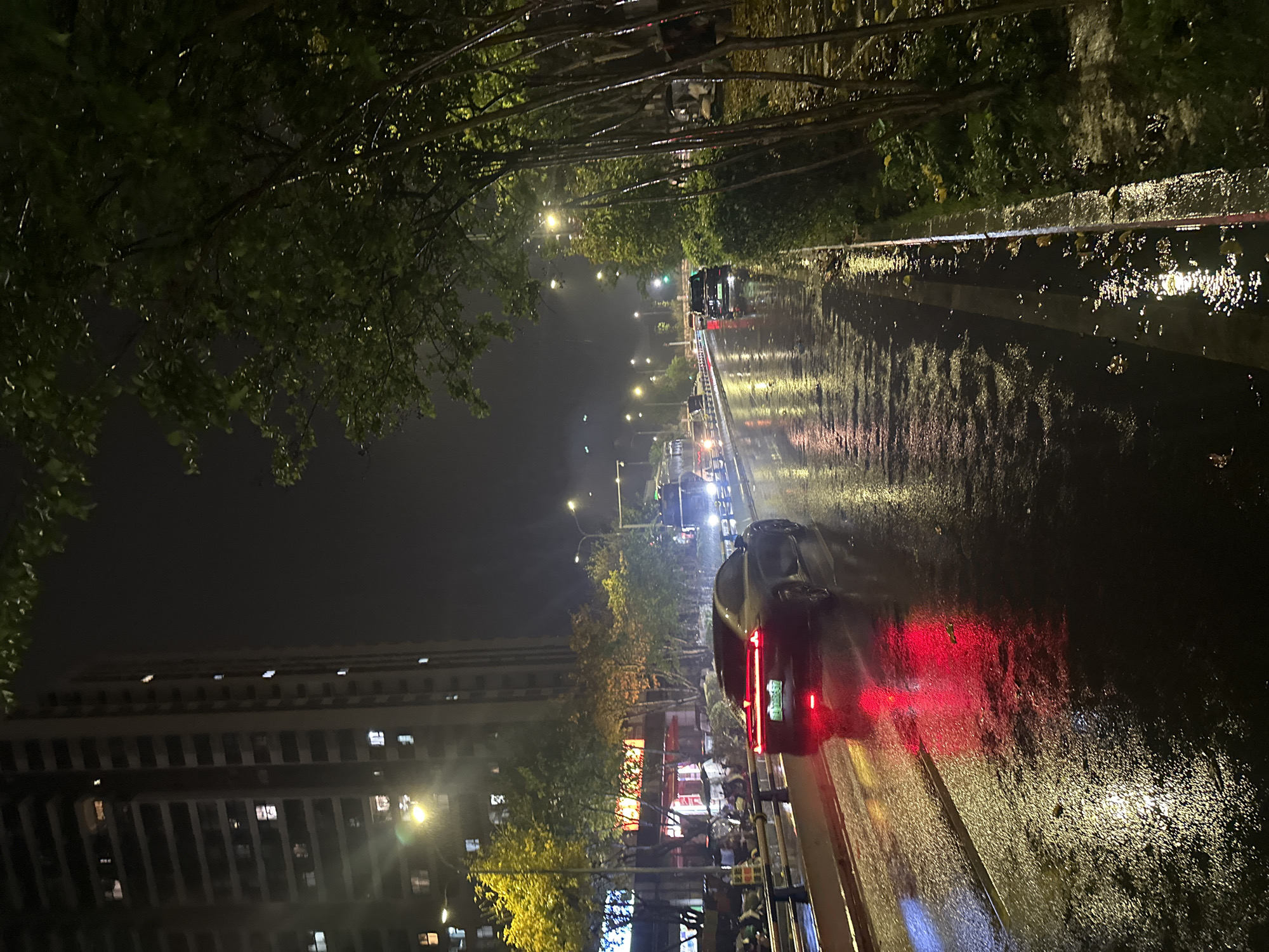 The image depicts a nighttime scene on a wet street in Chongqing High-tech Zone, China. Several vehicles are present on the road, including cars and a truck, with their headlights and taillights illuminated. The road surface reflects the city lights and vehicle lights, indicating recent or ongoing rain. Buildings line the side of the street, and trees are visible along the roadside. The lighting conditions suggest streetlights and building lights illuminate the area.
