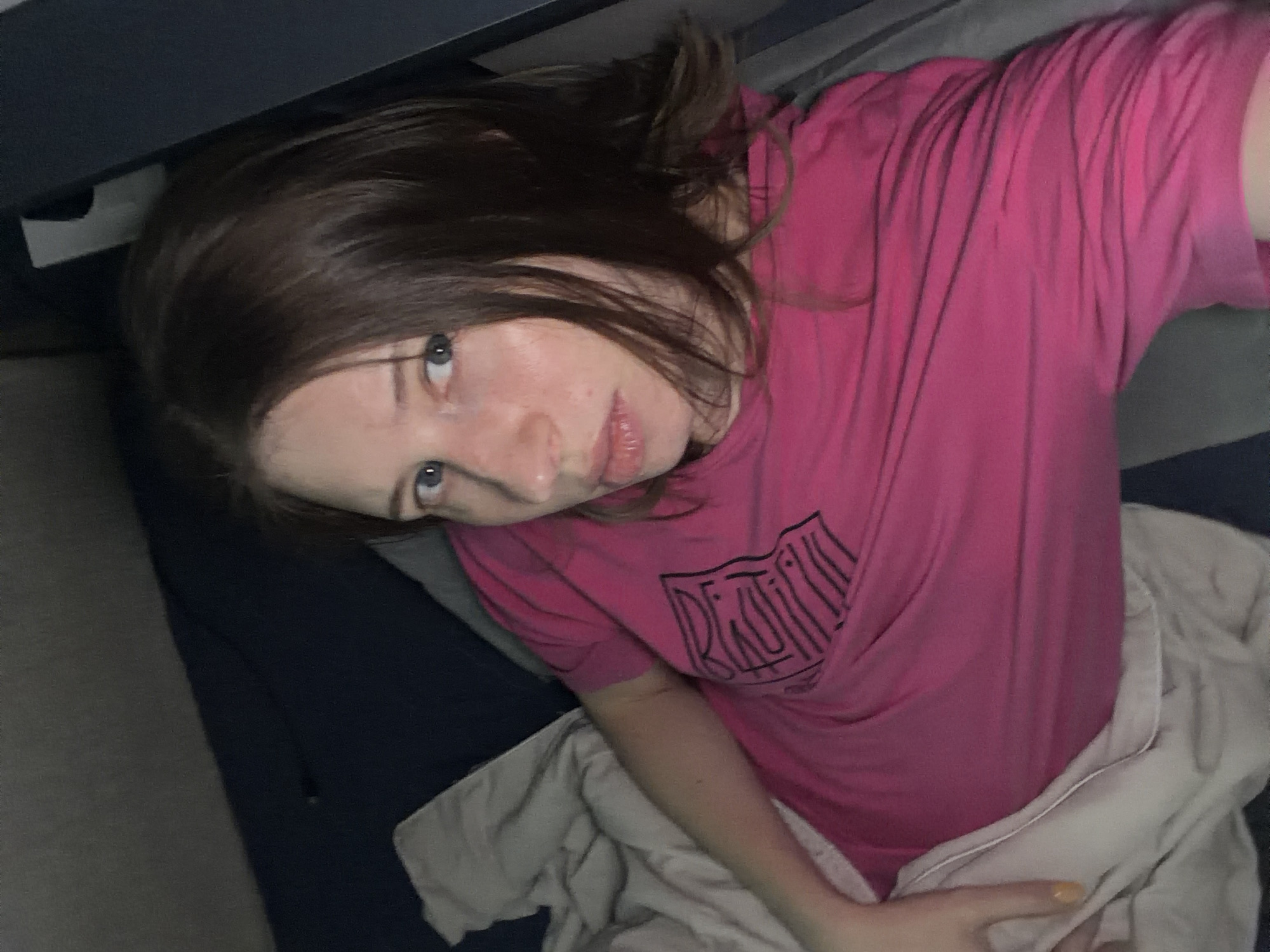 The image depicts a young woman lying down, covered partially by a blanket, wearing a bright pink t-shirt with a graphic design. She has light skin, brown hair, and blue eyes, with a neutral expression. The environment appears to be indoors, possibly a bedroom or a living space, with dark blue bedding visible and a dark grey surface to the right, suggesting furniture or a wall. The lighting is dim, casting shadows and creating a moody atmosphere. The provided information about the location being Dongducheon-si, South Korea, is not visually verifiable from the image itself.