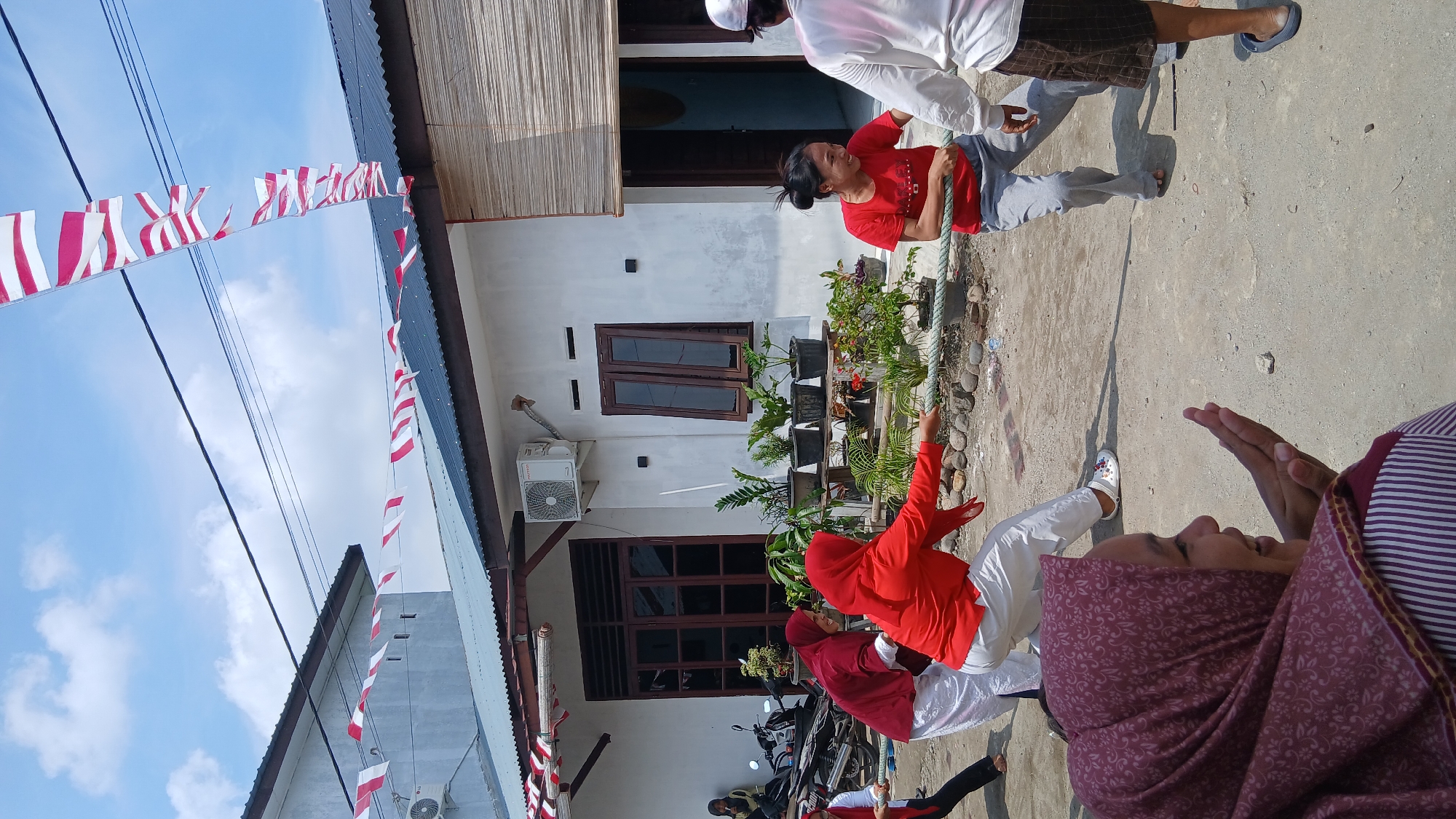 In Klambir Lima Kebun, Indonesia, a group of people are engaged in a tug-of-war competition. Three women are actively pulling a thick rope. One woman, wearing a red shirt and gray pants, is in the center, leaning back and pulling with all her might. To her left, another woman in a red headscarf and white pants is also pulling the rope, her body contorted with effort. On the far left, a third woman in a red headscarf and white pants is participating in the pull. In the foreground, a young woman wearing a patterned purple headscarf and a striped shirt looks towards the competition, her hands raised in an expressive gesture. The event appears to be taking place outdoors on a dirt path, with a white house in the background. The house has a dark-tiled roof and windows with dark frames. Several potted plants are arranged in front of the house. Red and white Indonesian flags are strung across overhead wires, indicating a possible celebration or national event. A motorcycle is parked to the left of the house. The sky is blue with scattered clouds.