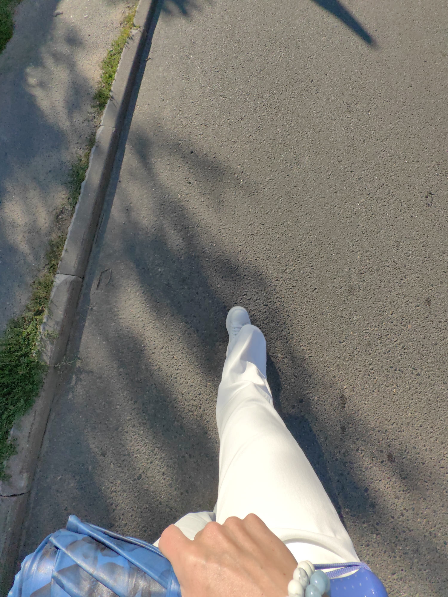 An over-the-shoulder shot shows a person walking on a paved surface, likely a sidewalk or road, with grass and a curb to the left. The person is wearing white pants and white shoes. They are carrying a blue bag with a black pattern in their left hand, which is resting on their lap. A beaded bracelet with blue and grey beads is visible on their right wrist. The ground is partially covered in shadows, suggesting it is daytime with sunlight filtering through. The location is identified as Fanipal, Belarus.