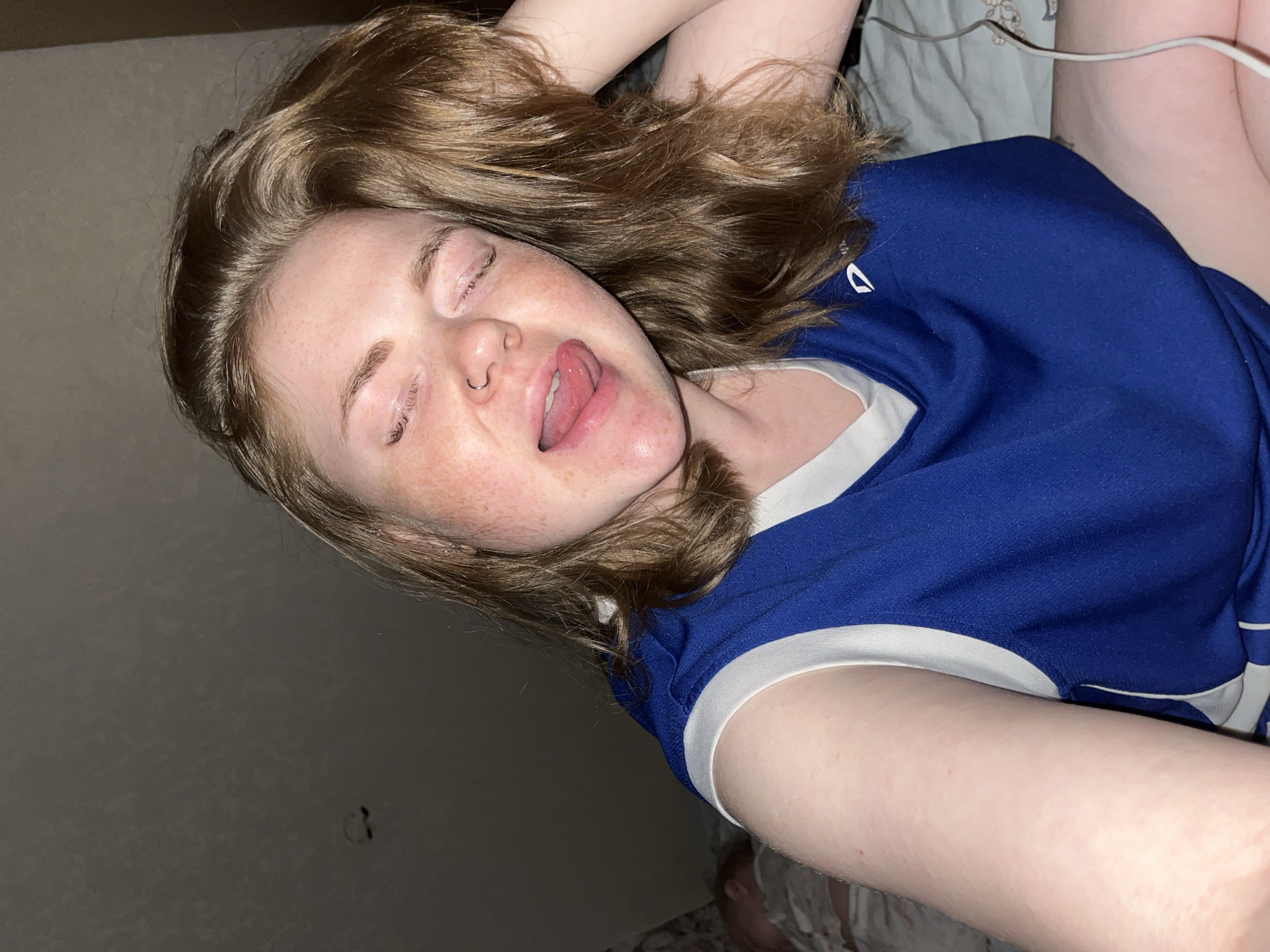 A young person with light skin and freckles is lying down, possibly on a bed. They have medium-length brown hair, closed eyes, and are sticking their tongue out. They are wearing a blue and white sports jersey. A silver nose ring is visible. The location is not identifiable as Chaikovsky, Russia, and is instead a poorly lit room with a wall visible in the background. There is a power cable visible behind the person.