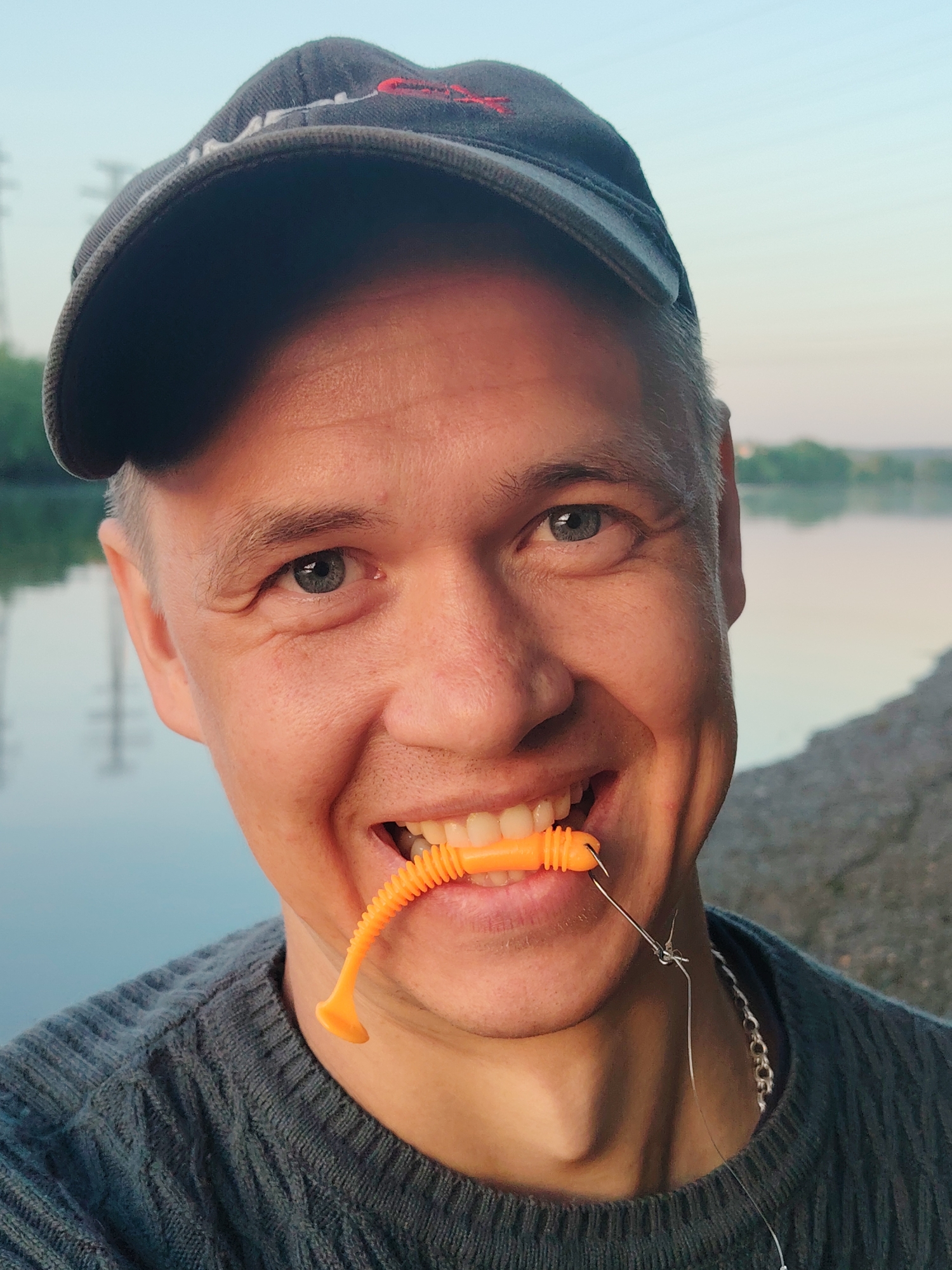 A man in a grey sweater and a dark baseball cap is smiling at the camera while holding an orange fishing lure in his mouth. The fishing lure has a hook attached to it with a fishing line, and a metal chain is also visible around his neck. He stands by a riverbank; in the background, a river can be seen, with trees visible on the opposite bank and some power lines visible in the distance. The image was taken in Chaikovsky, Russia.