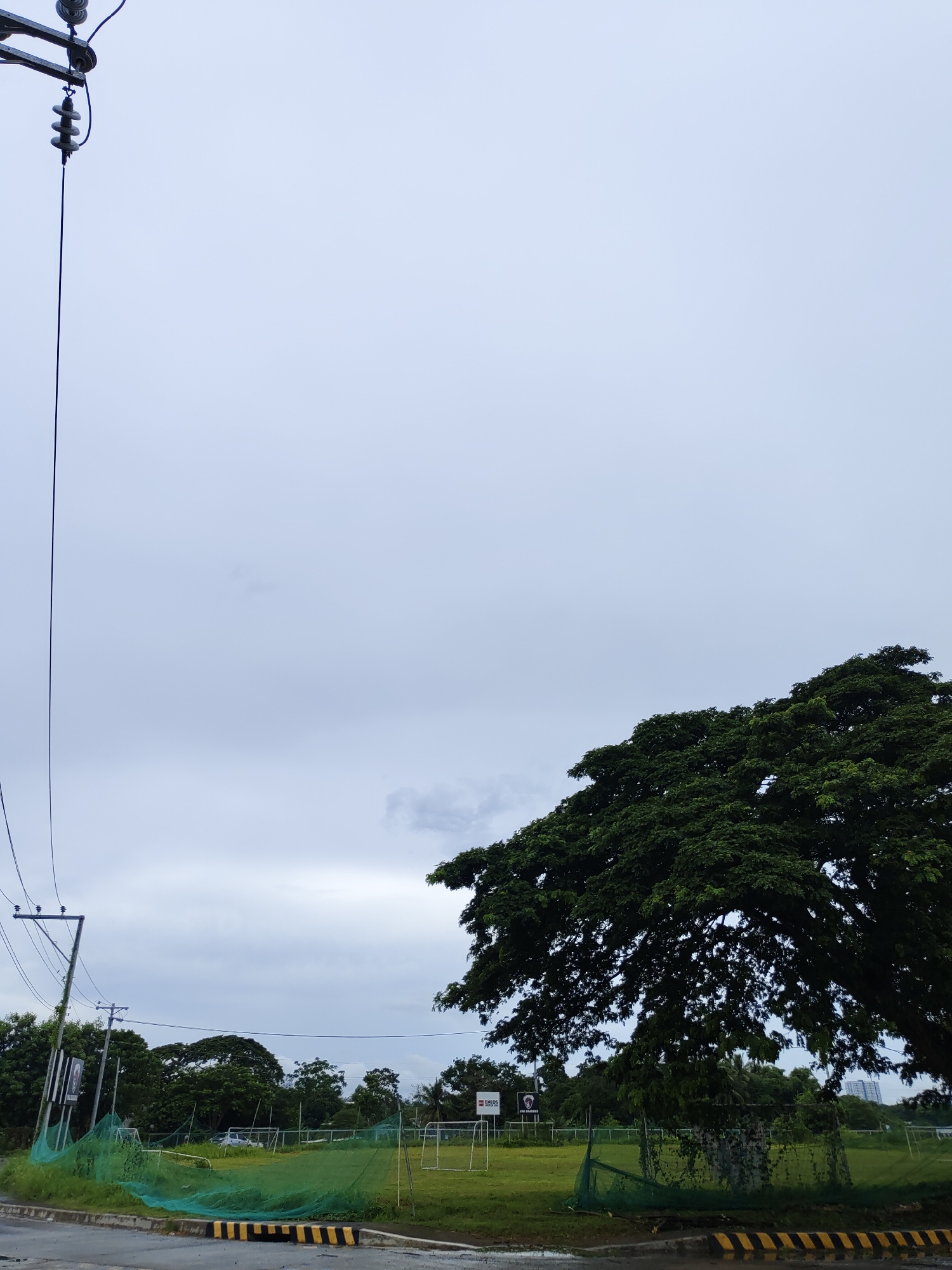 The image depicts an overcast sky over an outdoor sports field in Davao City, Philippines. A large, leafy tree dominates the right side of the frame, its branches spreading wide. In the foreground, a street curb with black and yellow warning stripes is visible, separating the road from a grassy area enclosed by a green safety net. Power lines and utility poles are present on the left side of the image, with a tall pole supporting multiple wires. A soccer or football field with goalposts can be seen in the background, along with some distant buildings and other trees. The overall scene suggests an urban or semi-urban outdoor recreational area under a cloudy sky.