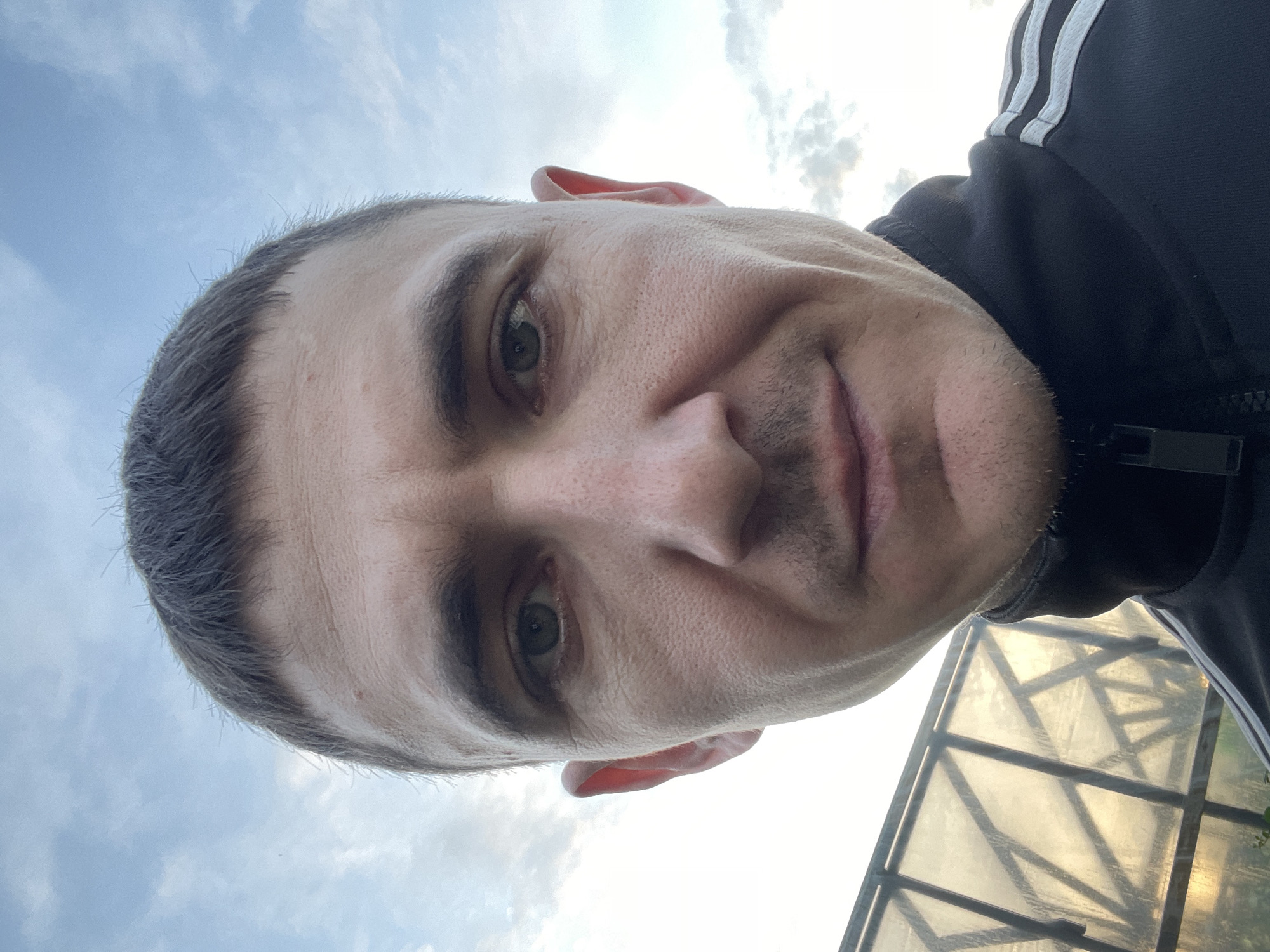 The image is a close-up, low-angle selfie of a man's face and upper torso. The man has short, dark hair, light skin, and is looking directly at the camera with a neutral expression. He is wearing a black zippered jacket with white stripes on the sleeves, which is characteristic of Adidas sportswear. In the background, to the left of the man, is a greenhouse structure made of glass or plastic panels supported by a metal frame. The sky above is cloudy and blue, suggesting an outdoor setting. The overall lighting indicates it may be late afternoon or early evening. The image does not contain any elements that specifically identify the location as Laishevo, Russia, or any other city.