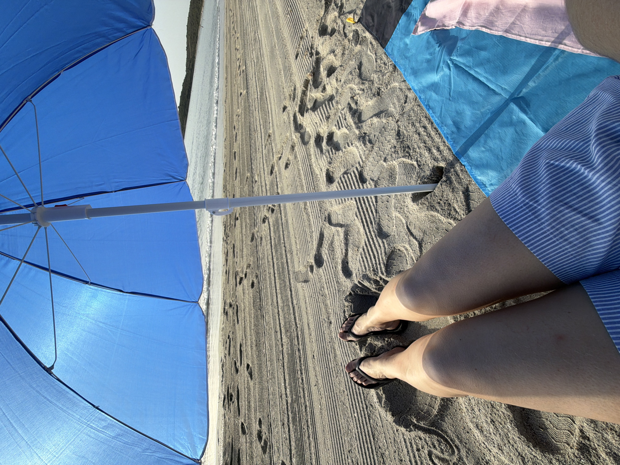 A first-person perspective shot of a person sitting on a sandy beach under a large blue umbrella. The sand is packed and has numerous footprints and rake marks. The ocean and a distant shoreline with vegetation are visible in the background. The person is wearing black flip-flops and blue and white striped shorts. A blue beach towel and a pink cloth are laid out on the sand next to the person. The scene is bathed in sunlight, casting shadows. This appears to be a relaxed setting on a beach, possibly in the city of Olbia, Italy, given the typical coastal scenery.