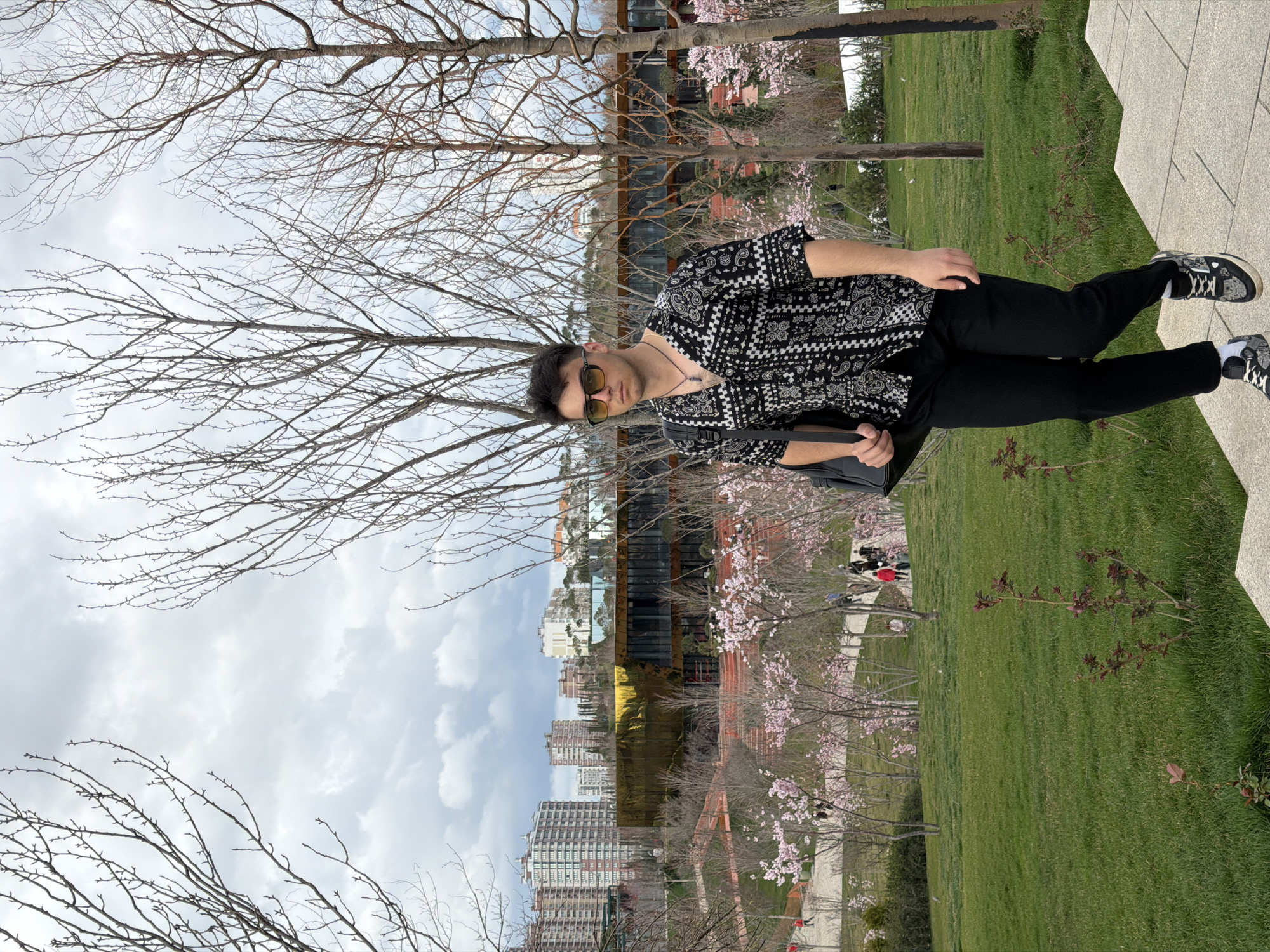 A medium shot depicts a young man standing in a park. He is facing the camera, wearing a black and white patterned short-sleeved shirt, black pants, and black and white sneakers. He has sunglasses on and a dark bag slung over his shoulder. The background includes trees, some blossoming with pink flowers, a modern building with gold mirrored windows, and city buildings in the distance under a cloudy sky. A paved area and grass surround him. Based on the architecture and environment, this may be near the FC Krasnodar Stadium in Krasnodar, Russia.