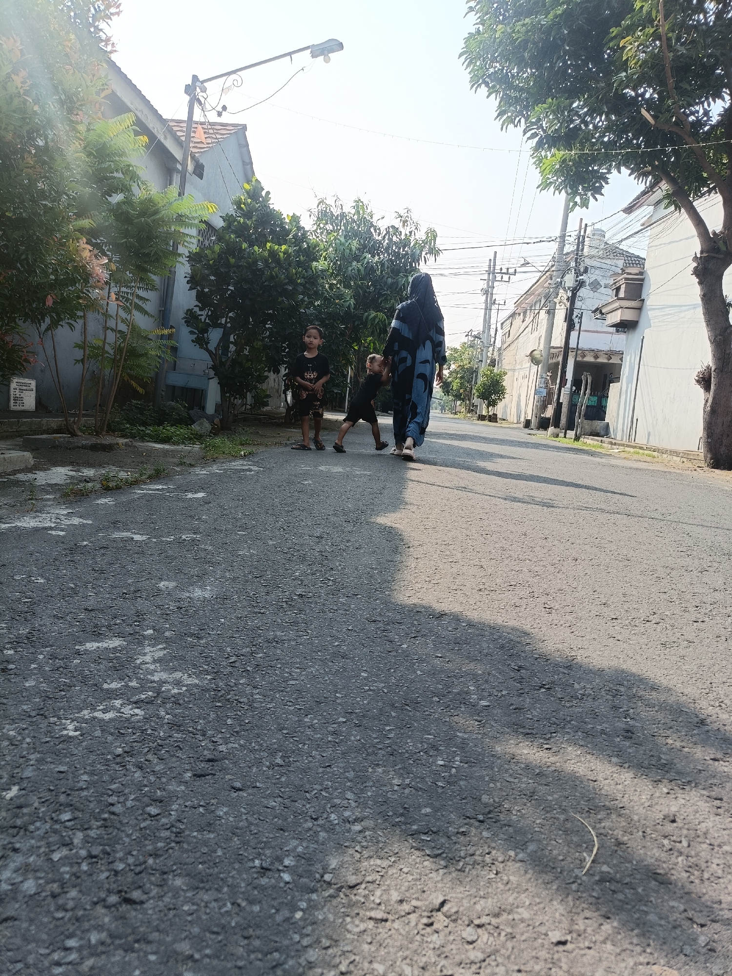 The image shows a street scene in Kediri City, Indonesia, on a sunny day. In the foreground, a paved road with scattered lighter patches and shadows from trees occupies most of the frame. In the mid-ground, a woman wearing a dark hijab and a patterned blue and black outfit is walking away from the camera, holding the hand of a young child in a black t-shirt and shorts. To the left of them, another young child in a black t-shirt and patterned shorts stands facing the camera. The children appear to be a boy and a girl, respectively. Behind them, houses with light-colored walls and tiled roofs line the street. Trees with green foliage are visible on both sides of the street and in front of the houses. A street light is mounted on a pole on the left side of the road, and power lines are strung across the sky. The overall atmosphere is one of a quiet residential street.