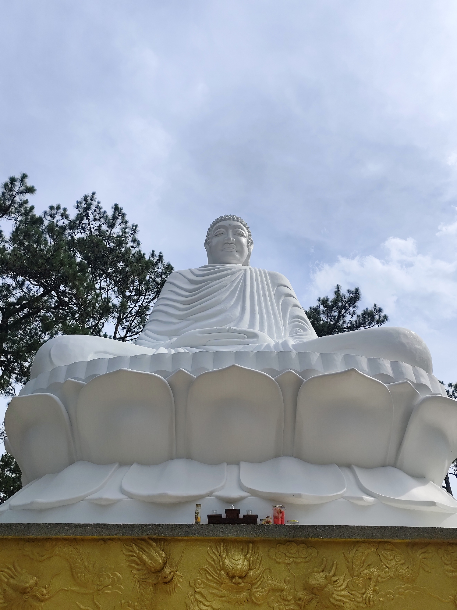 The image depicts a large, white Buddha statue seated in a lotus position. The statue is placed on a platform with golden dragon carvings. There are small offerings placed in front of the statue. Green trees are visible in the background, and the sky is overcast. The location is the city of Da Lat, Vietnam.