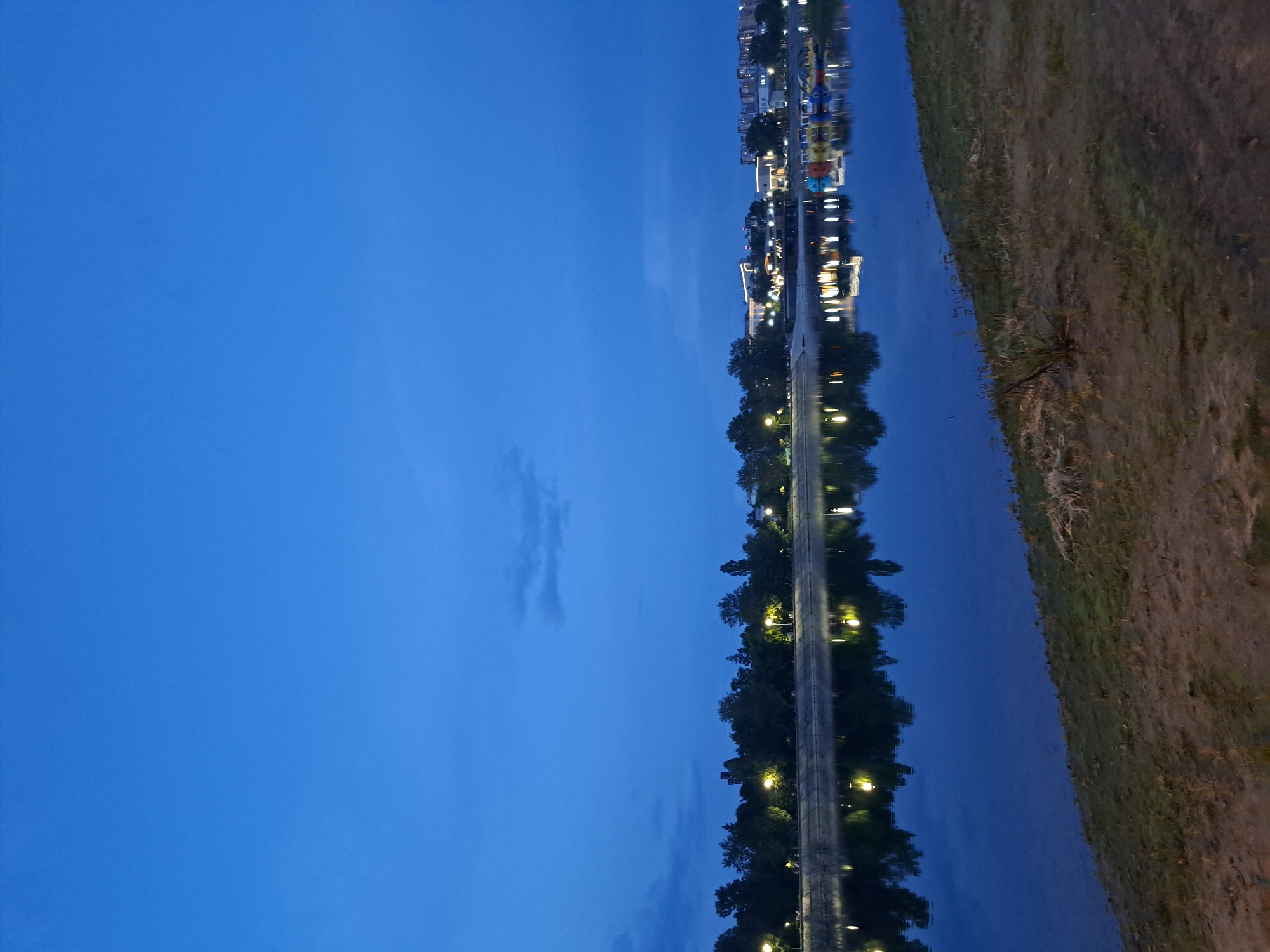 The image depicts a tranquil twilight scene along a body of water, likely a river or lake. The sky is a deep, vibrant blue, with faint wisps of clouds. A line of trees with glowing streetlights can be seen along the far bank, casting reflections onto the still water. Beyond the trees, buildings and structures are visible, illuminated by artificial lights, suggesting an urban or semi-urban setting. On the water, there are several colorful paddle boats, adding a touch of vibrancy to the otherwise muted tones of the evening. The foreground consists of a grassy and sandy embankment. This scene is consistent with the embankment of the Angara River in Irkutsk, Russia, particularly the area known for its recreational activities and promenade. No people are discernible in the image.