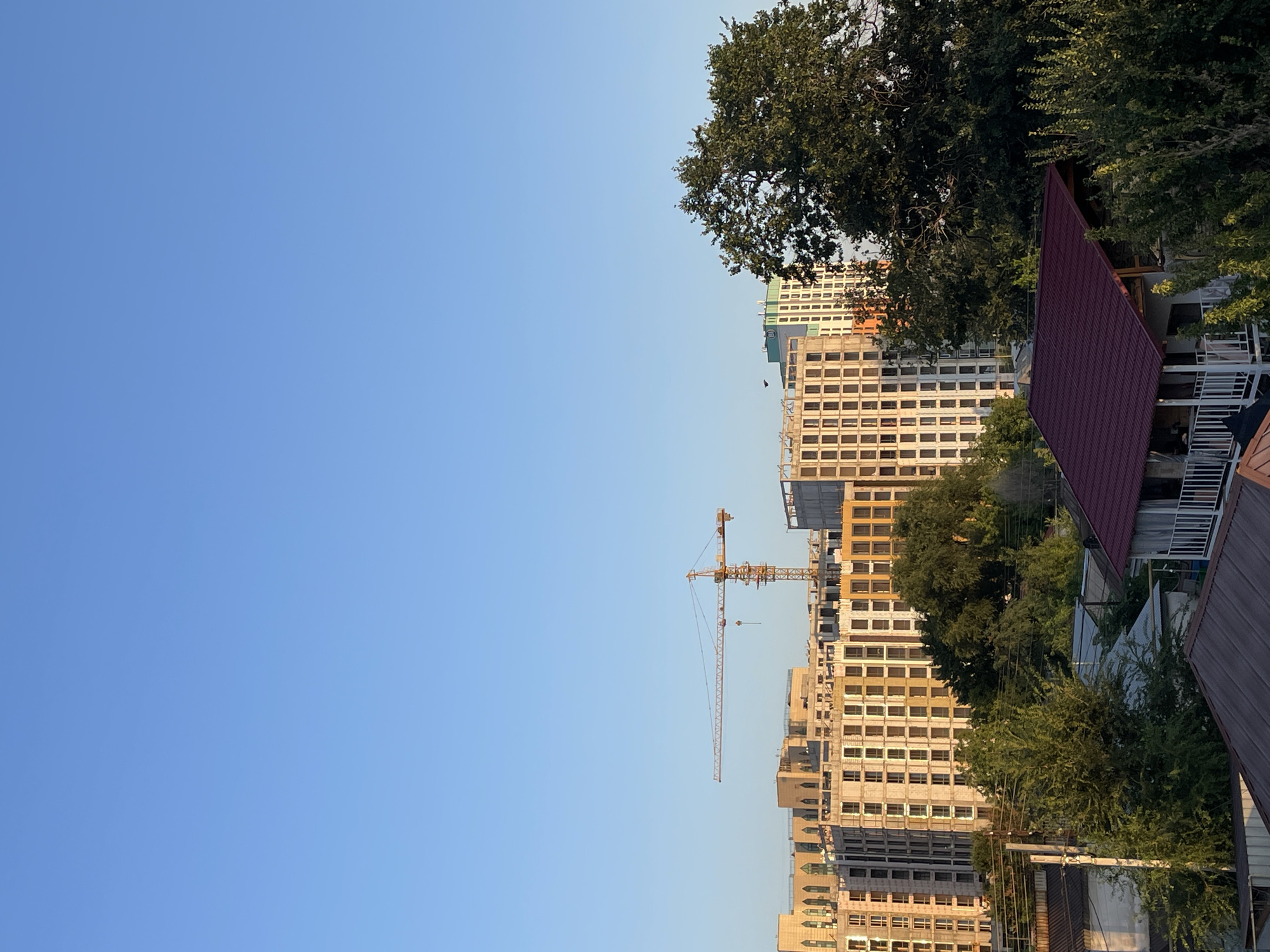 The image shows a city skyline in Almaty, Kazakhstan, under a clear blue sky. In the mid-ground, a construction site features several tall buildings in various stages of completion. A yellow tower crane is visible, positioned between two of the structures, suggesting ongoing construction work. The buildings display a mix of architectural styles, with some featuring arched windows and others a more modern, grid-like facade.
In the foreground, a dense canopy of green trees partially obscures lower structures, including houses with dark red roofs. One house has a covered porch with a white railing. The presence of power lines strung between poles adds to the urban setting. The lighting suggests it is late afternoon or early evening, with the sun casting a warm glow on the buildings. The scene captures a blend of new development and established residential areas within the city.