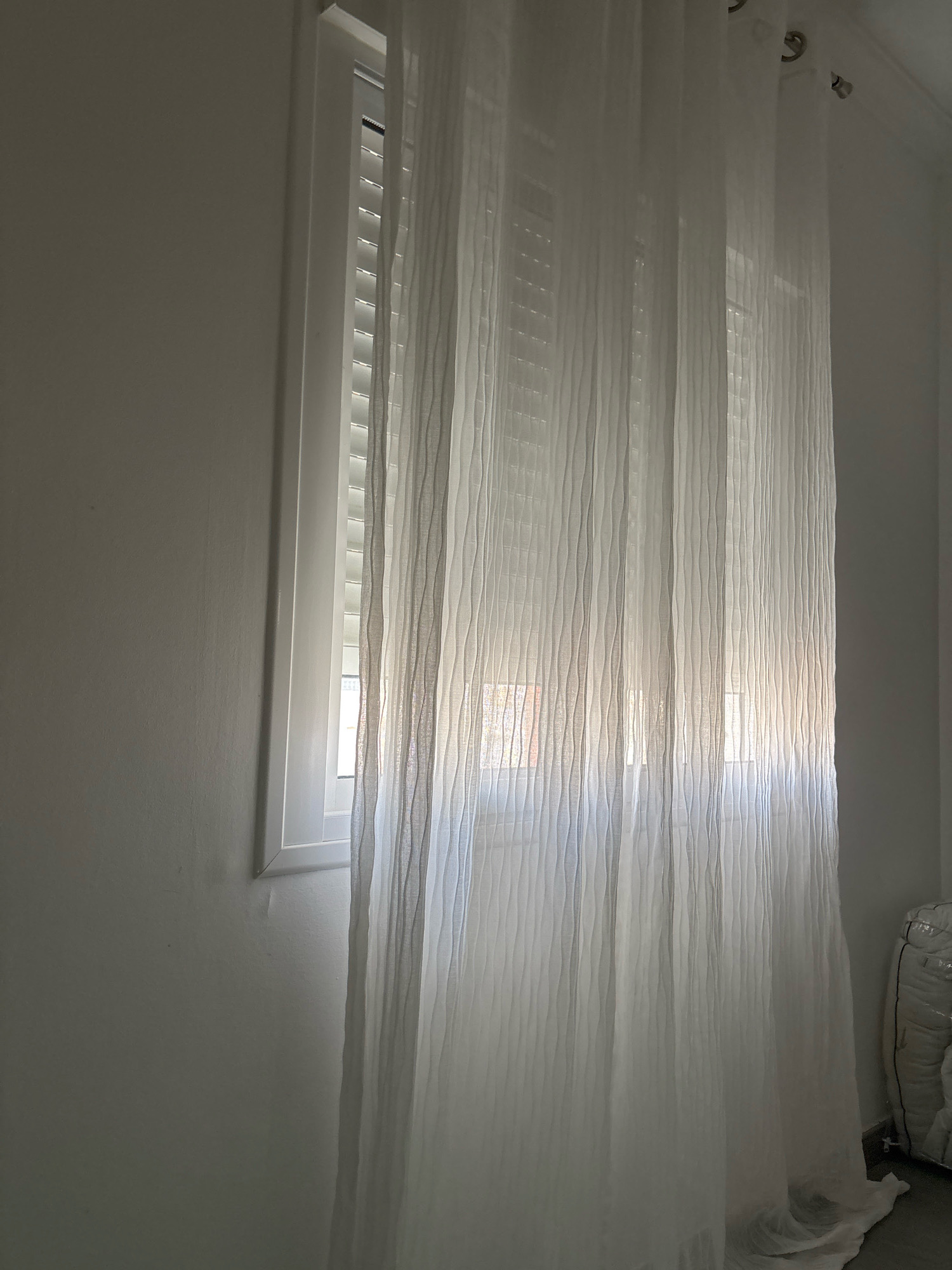 The image shows a white, sheer curtain with vertical pleats hanging in front of a window. The window is framed by a white molding and has blinds. Sunlight is filtering through the curtain, creating a soft glow. To the right of the curtain, a stack of white, bagged items is visible against a plain wall. The overall scene suggests an indoor setting, possibly a residential or commercial space, with natural light illuminating the area. There is no discernible indication of the city of Málaga, Spain.