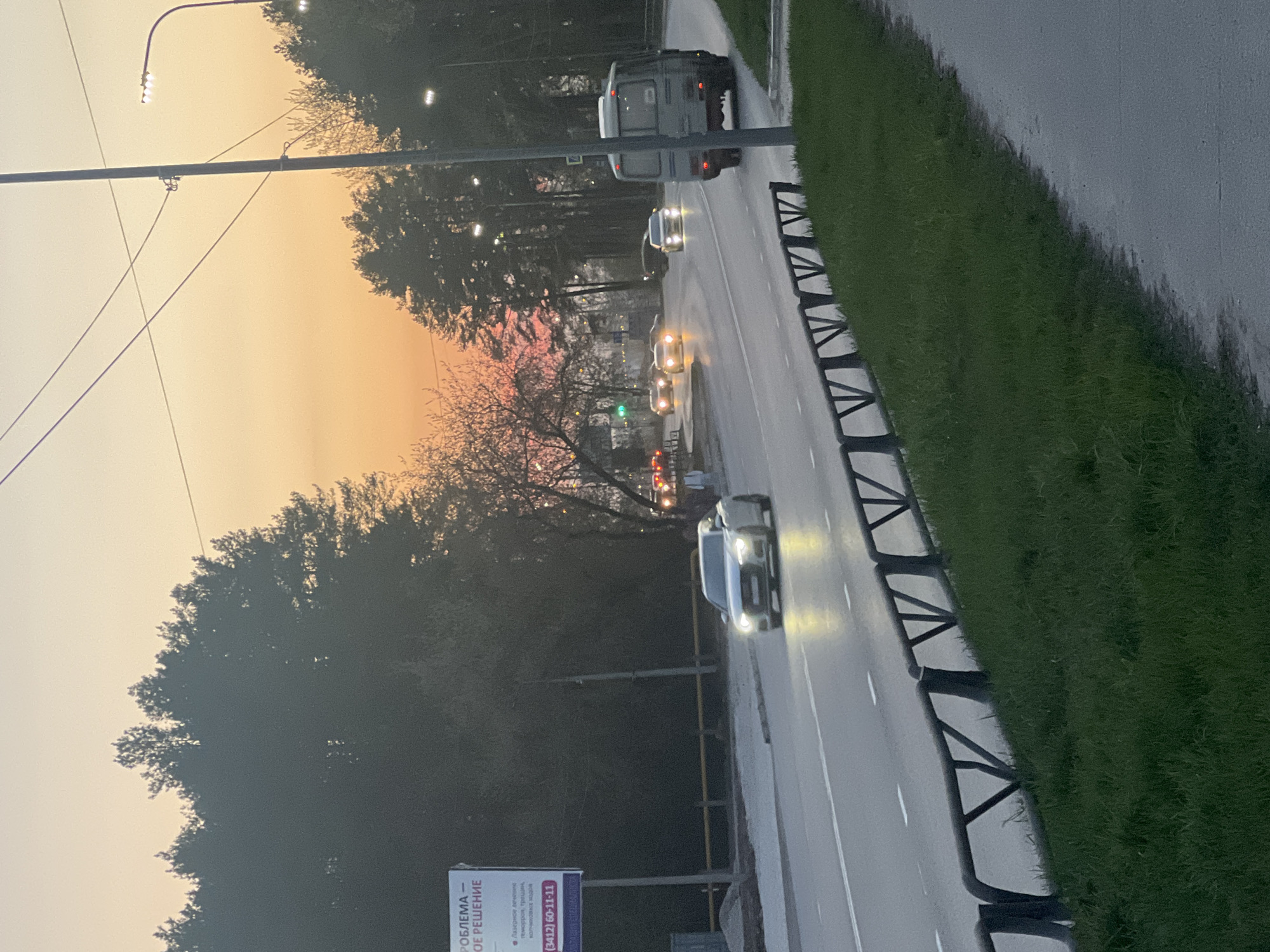 The image captures a road in Chaikovsky, Russia, at twilight. The sky is a gradient of orange and peach. A line of vehicles with headlights on travels along the road, which is lined on one side with a green verge and a protective barrier. A billboard with Russian text is positioned near the road. Overhead, a streetlight extends over the road. Trees are visible in the background, silhouetted against the sky.